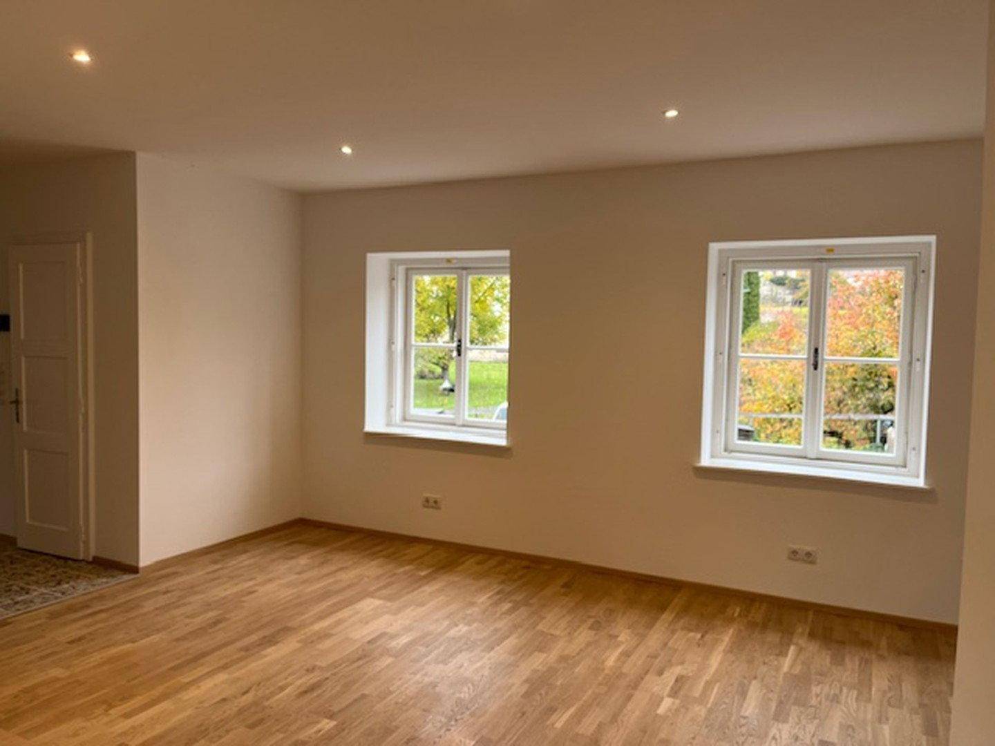 Heller Wohnraum mit Holzboden, weißen Wänden und zwei Fenstern mit Blick ins Grüne.