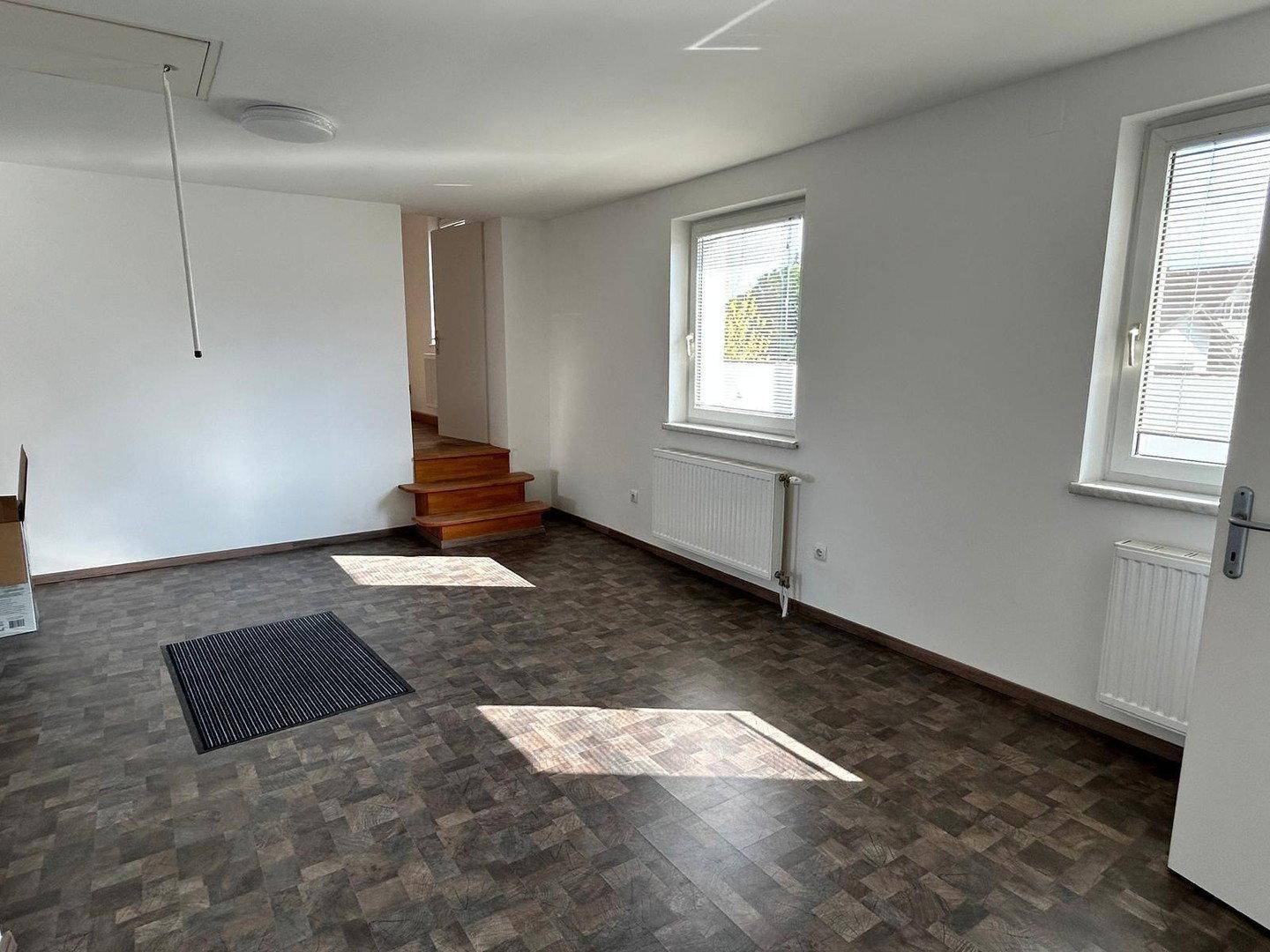 Geräumiges Wohnzimmer mit dunklem Fliesenboden, hellen Wänden und Fenstern mit Jalousien.