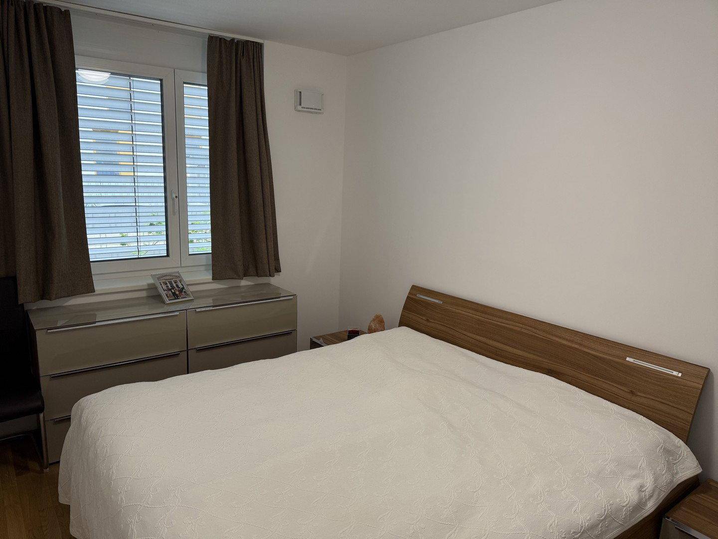 Schlafzimmer mit Doppelbett, Holzboden und Fenster mit braunen Vorhängen.