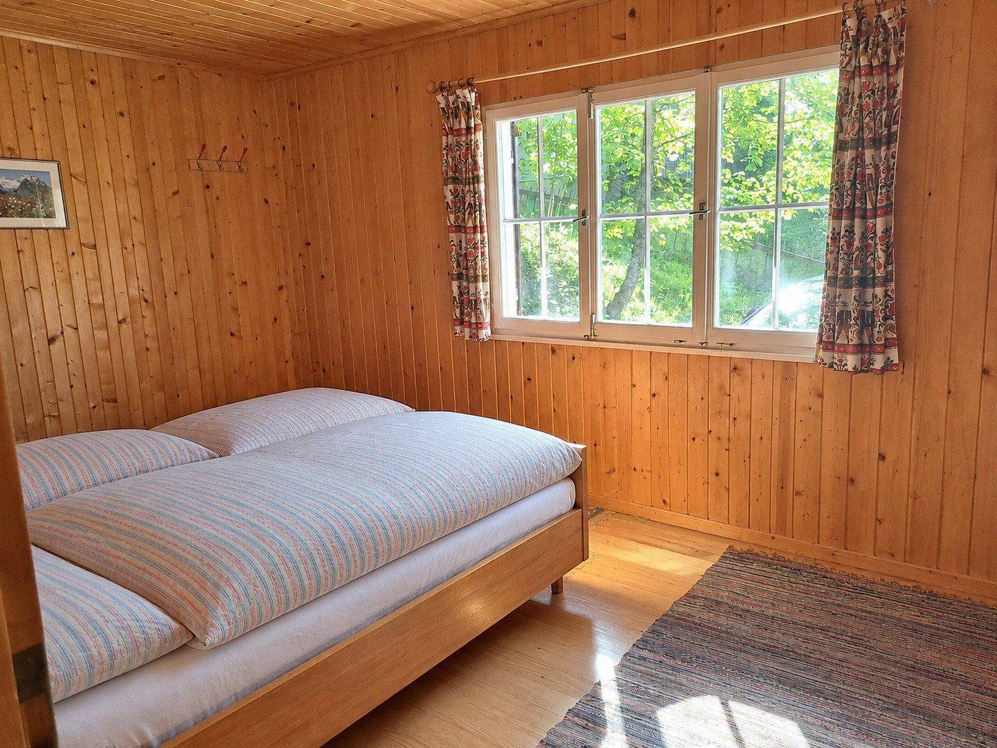 Helles Schlafzimmer mit Holzboden, Holzvertäfelung und zwei Einzelbetten, mit Blick ins Grüne.