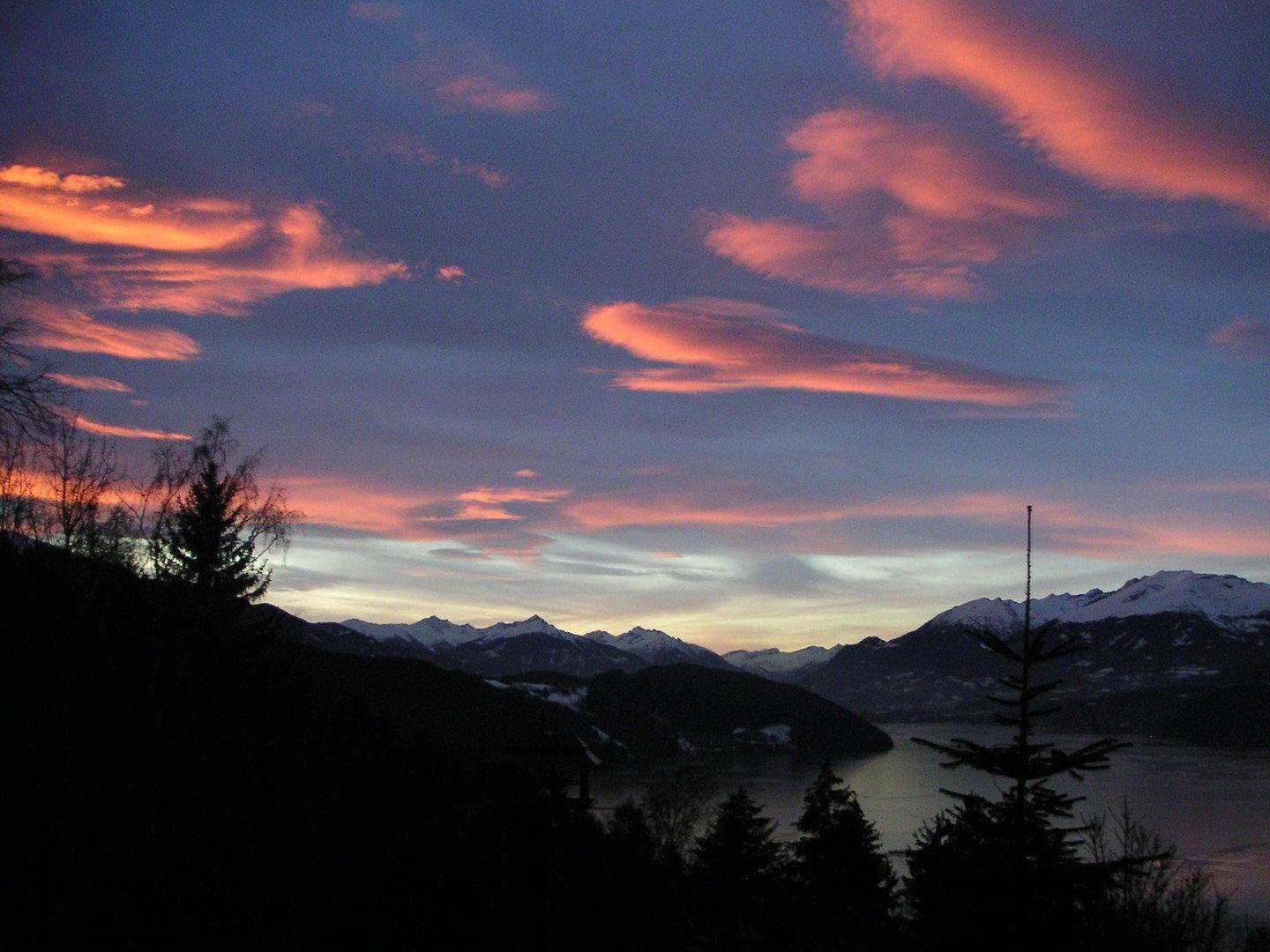 Atemberaubender Sonnenuntergang mit leuchtend orangefarbenen Wolken über einem See und schneebedeckten Bergen.