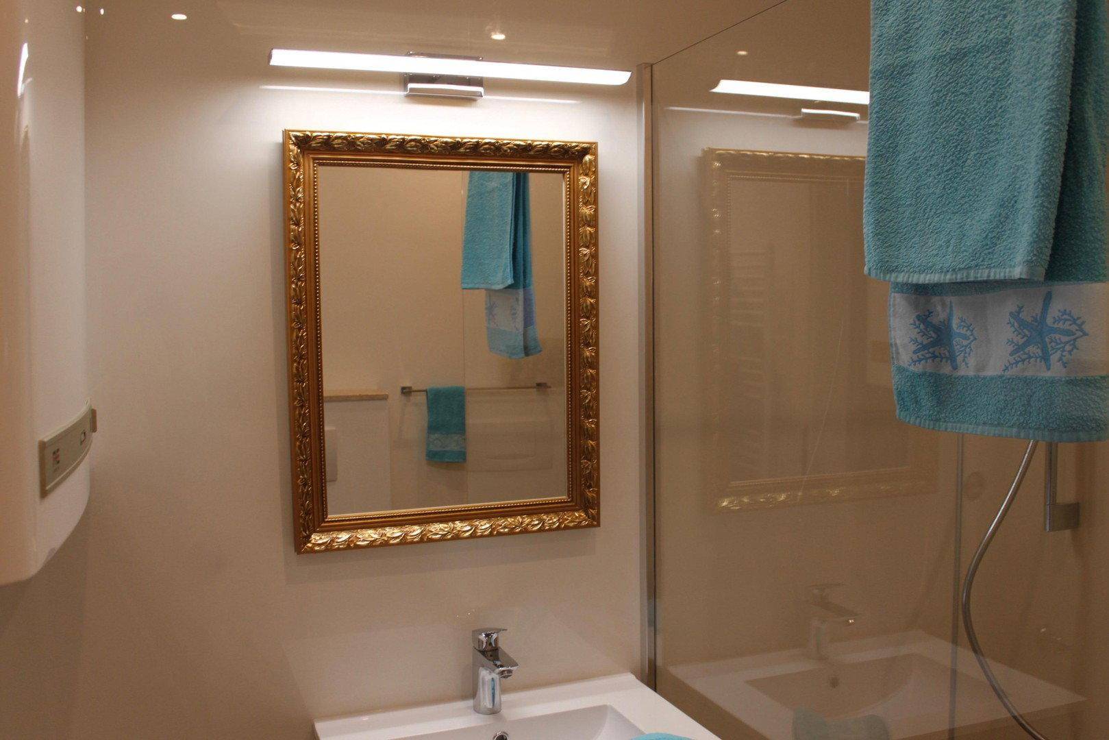 Helles Badezimmer mit Einzelwaschbecken, vergoldetem Spiegel und Duschbereich mit Glasabtrennung.