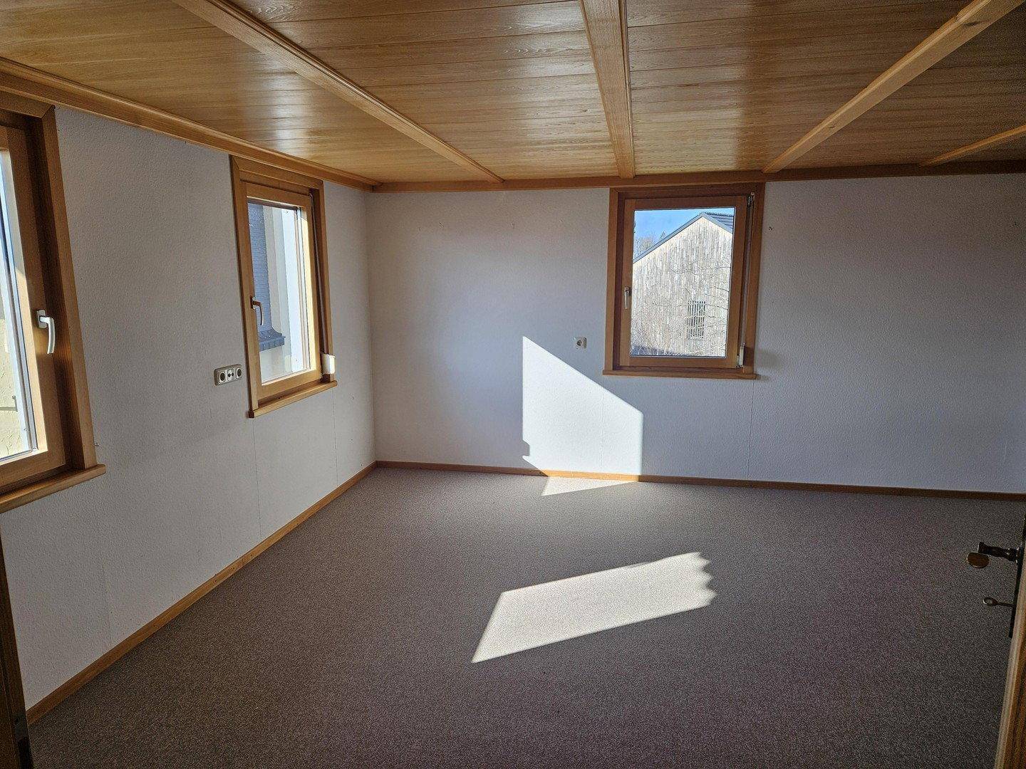 Helles Wohnzimmer mit Teppichboden, Holzdecke und mehreren Fenstern für Tageslicht.