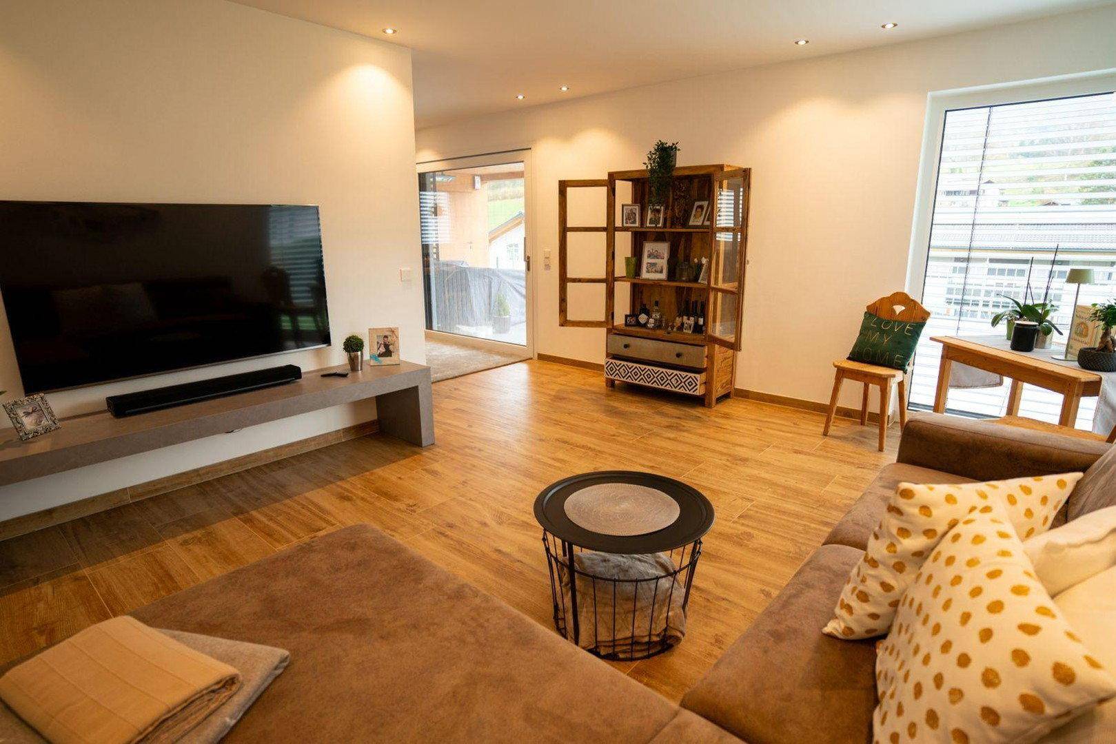 Heller Wohnbereich mit großem Fernseher, Holzboden und Zugang zu einem weiteren Raum.
