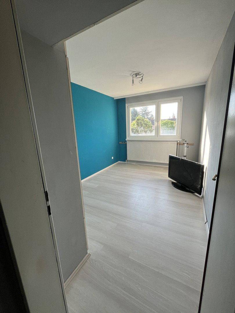 Helles Zimmer mit blau gestrichener Akzentwand, Laminatboden und großem Fenster.