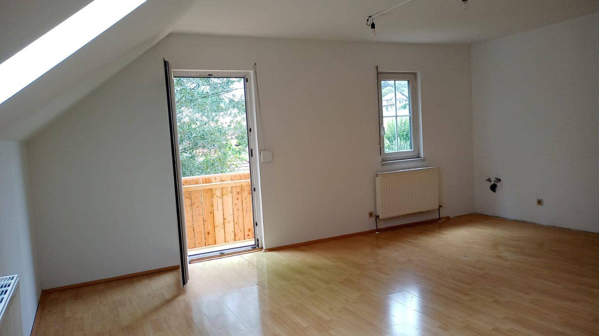 Heller Raum mit Dachschräge, Holzboden und Zugang zu einem kleinen Balkon, ideal als Wohn- oder Schlafzimmer.