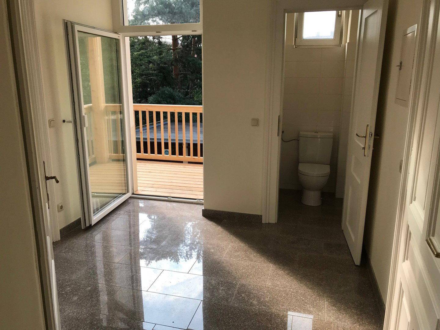 Heller Innenraum mit glänzendem Fliesenboden, Zugang zu einem Balkon und einer separaten Toilette.