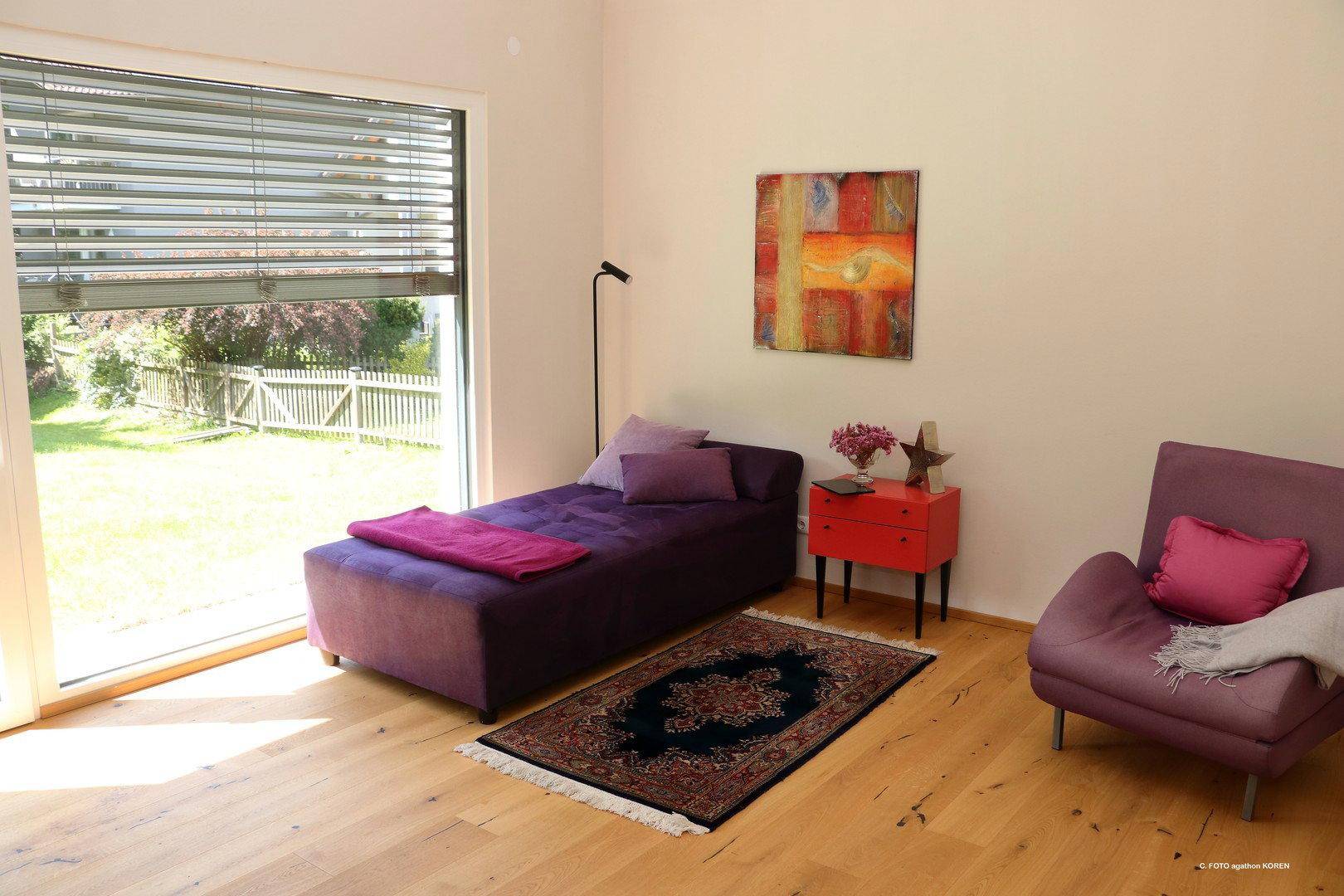 Gemütlicher Wohnbereich mit lila Chaiselongue, Holzboden und großen Fenstern zum Garten.