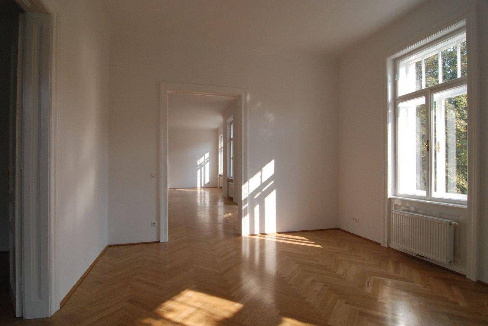 Helles, leeres Zimmer mit Fischgrätparkett und hohen Fenstern, die viel Sonnenlicht hereinlassen.