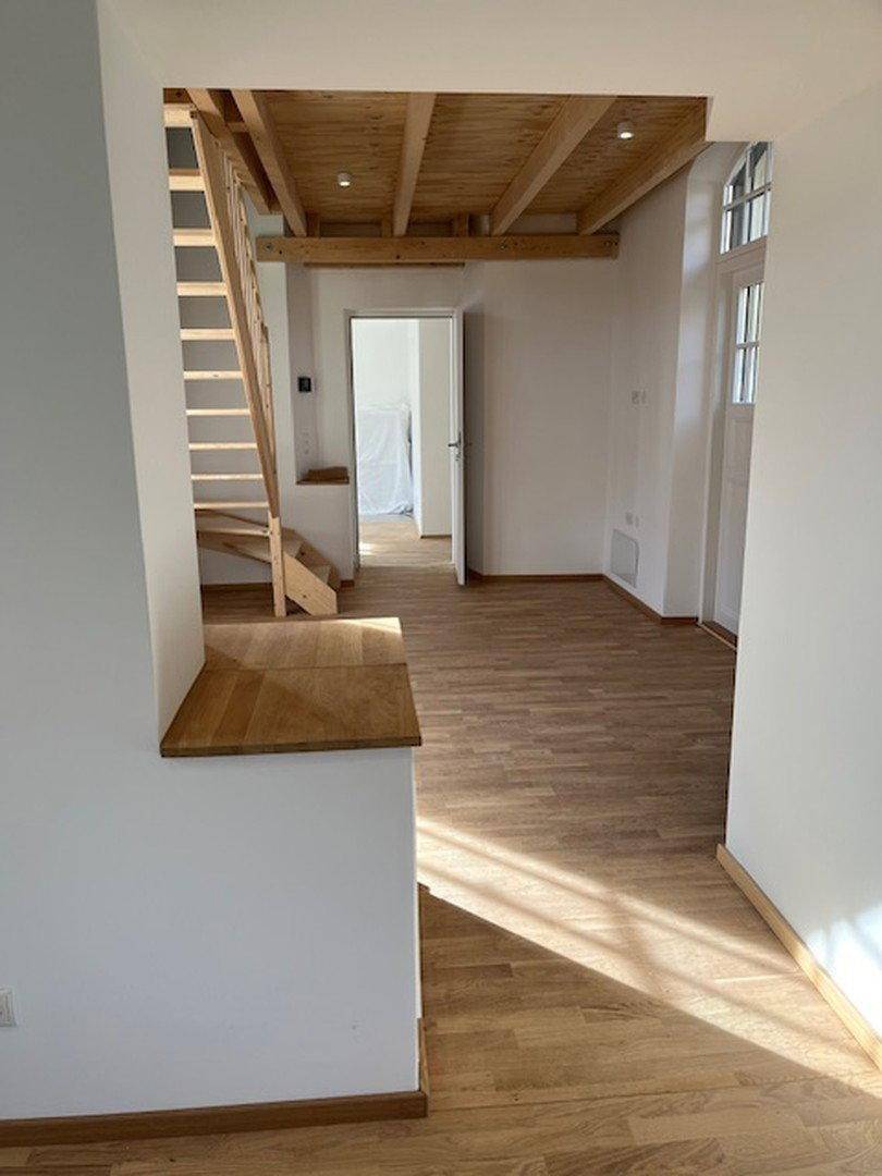 Heller Wohnbereich mit Holzboden, sichtbaren Holzbalken und einer Holztreppe.