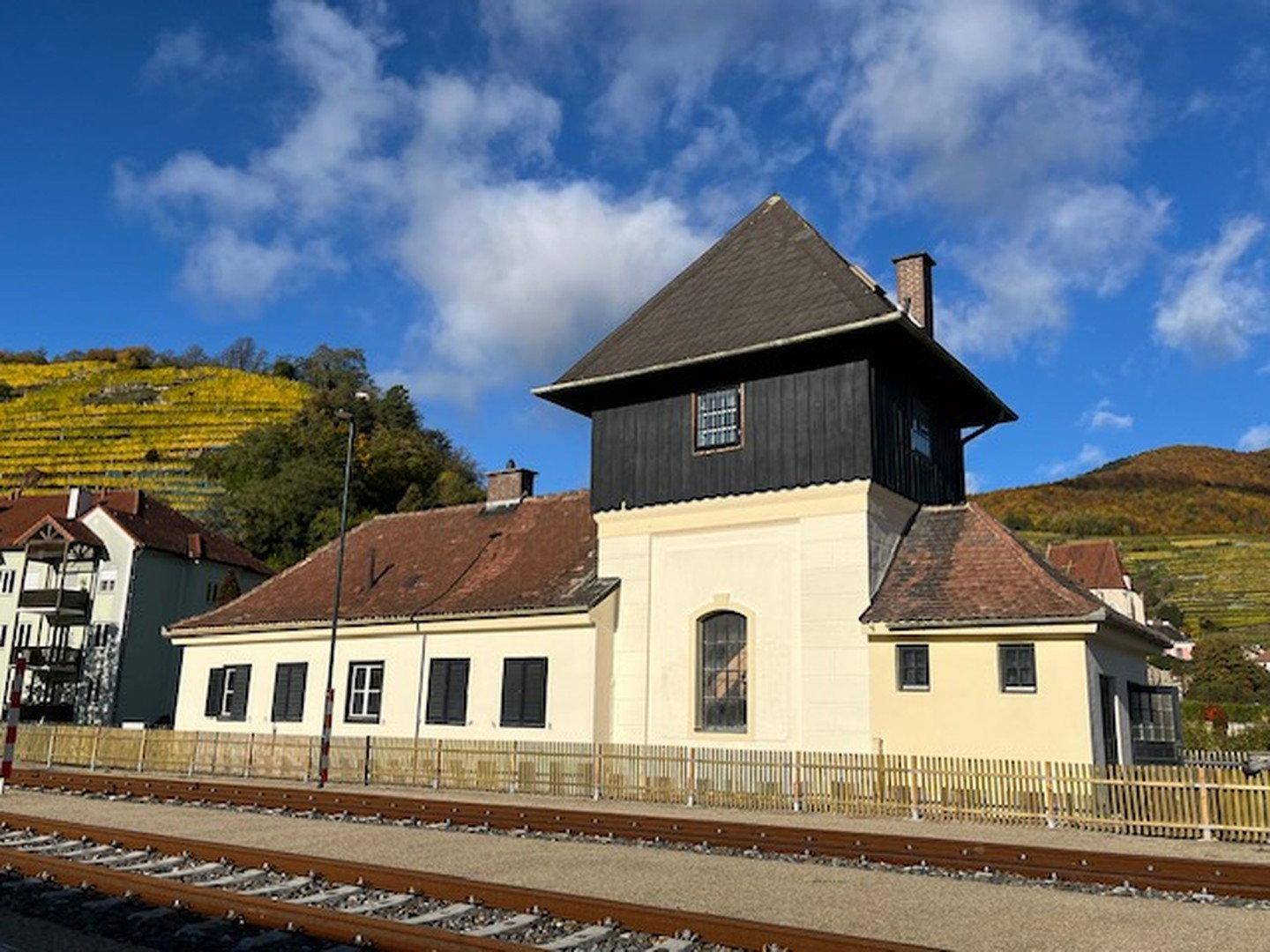 Historisches Gebäude neben Bahngleisen mit Weinbergen im Hintergrund unter blauem Himmel.
