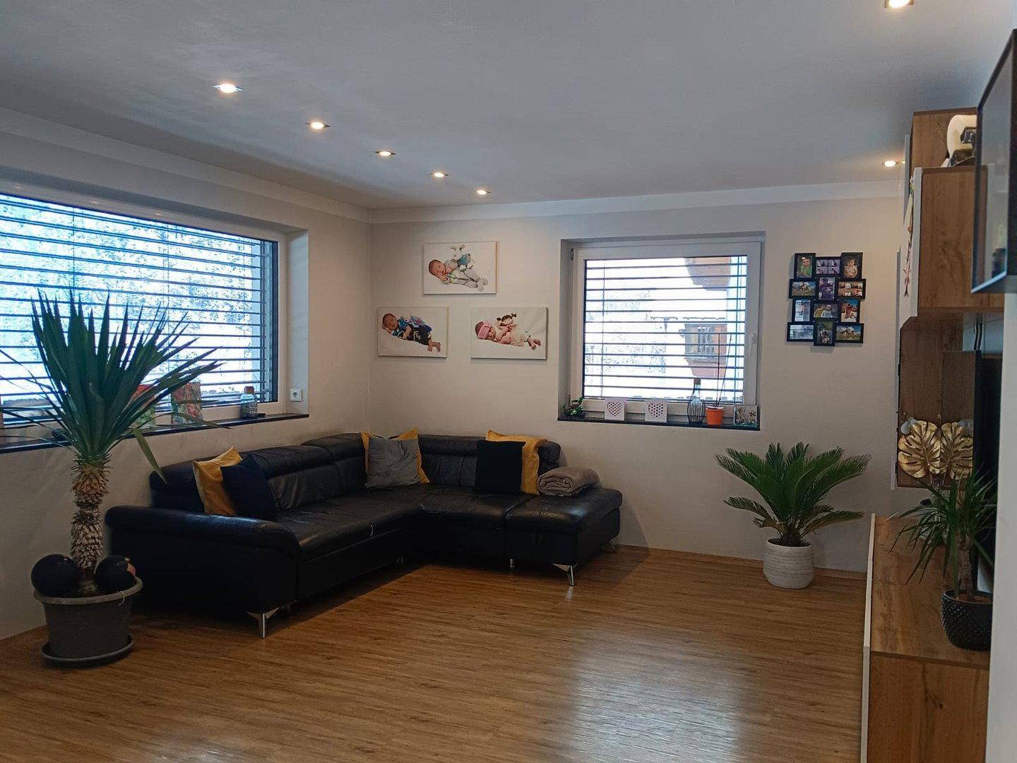 Gemütliches Wohnzimmer mit dunklem Ecksofa, Holzboden und großen Fenstern.