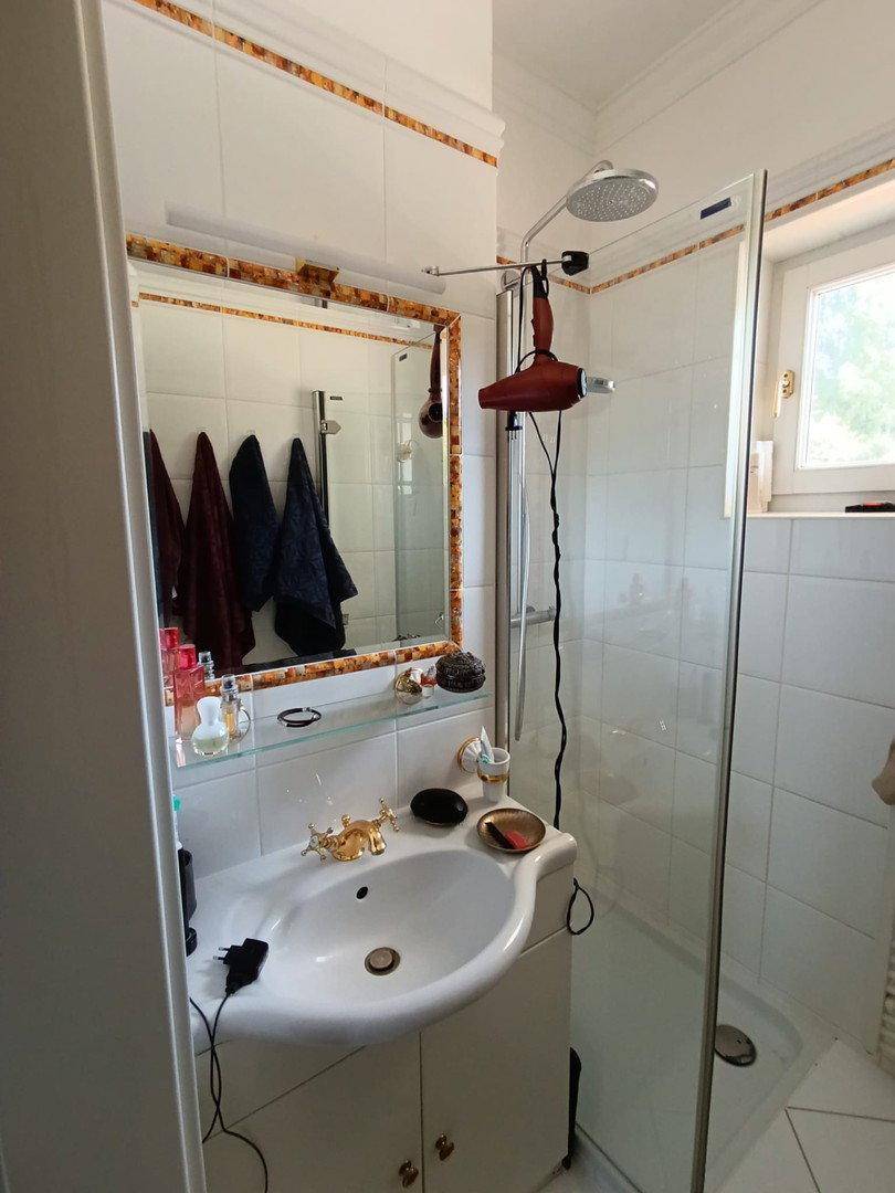 Kleines Badezimmer mit Dusche, Waschbecken, Spiegel und Fenster für Tageslicht.