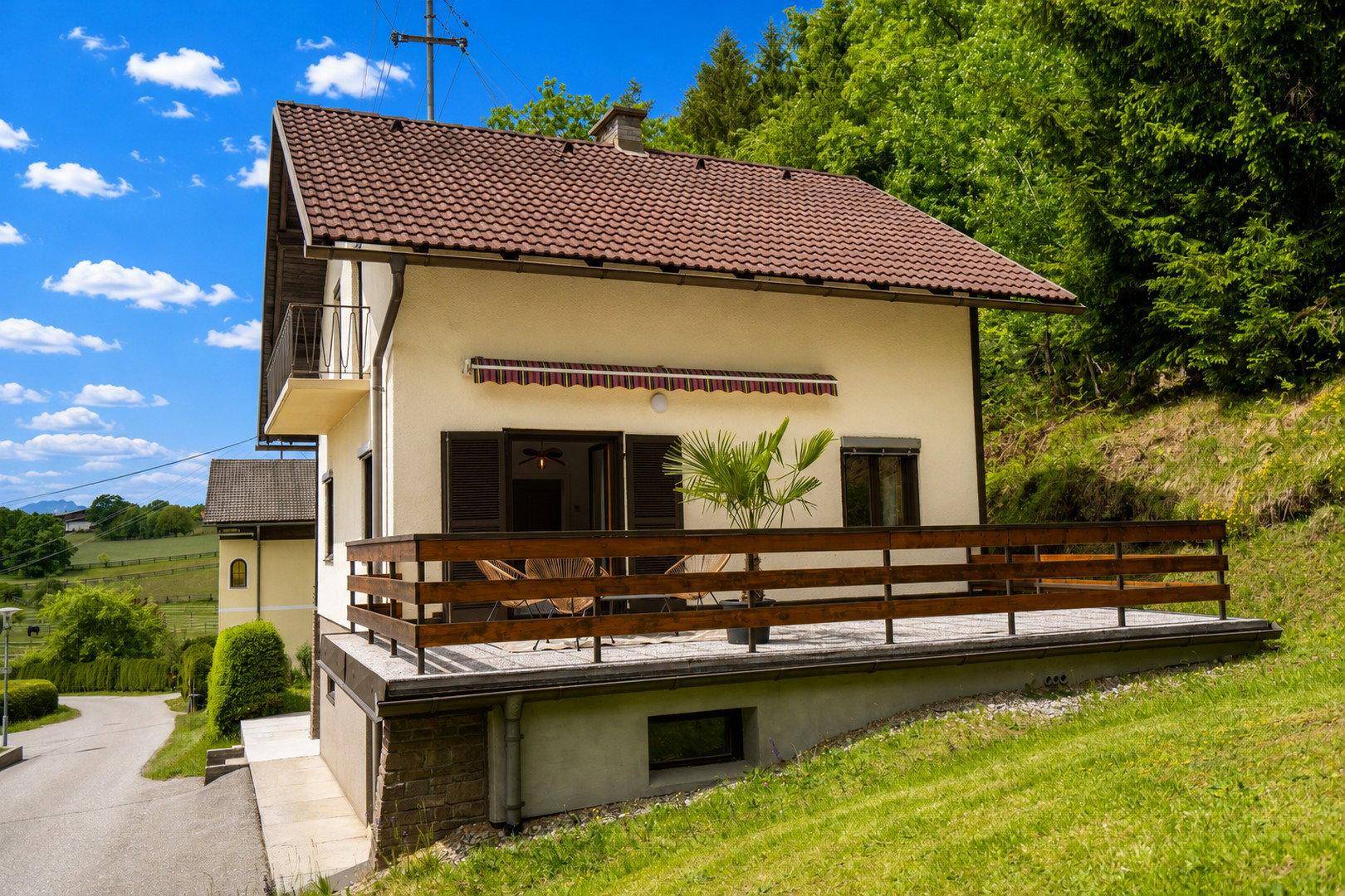 Haus mit großer Holzterrasse und Markise, umgeben von grüner Landschaft und Bäumen.