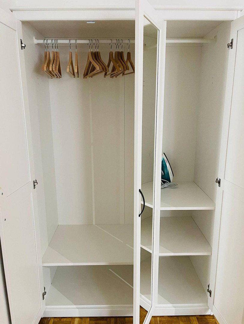 Offener weißer Kleiderschrank mit Kleiderstange, Regalböden und Bügeleisen.