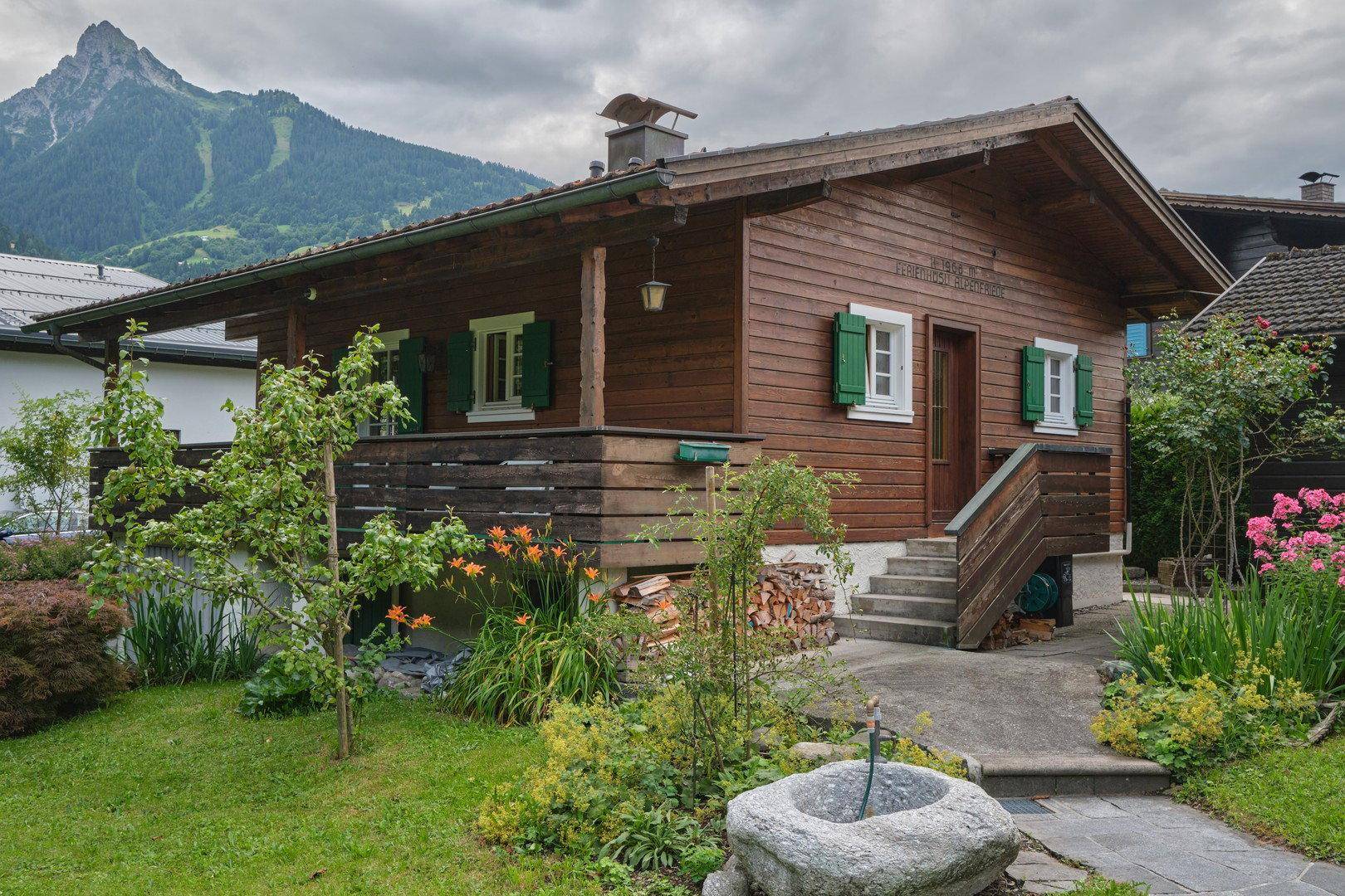 Rustikales Holzhaus mit grünen Fensterläden und einem gepflegten Garten vor Bergkulisse.