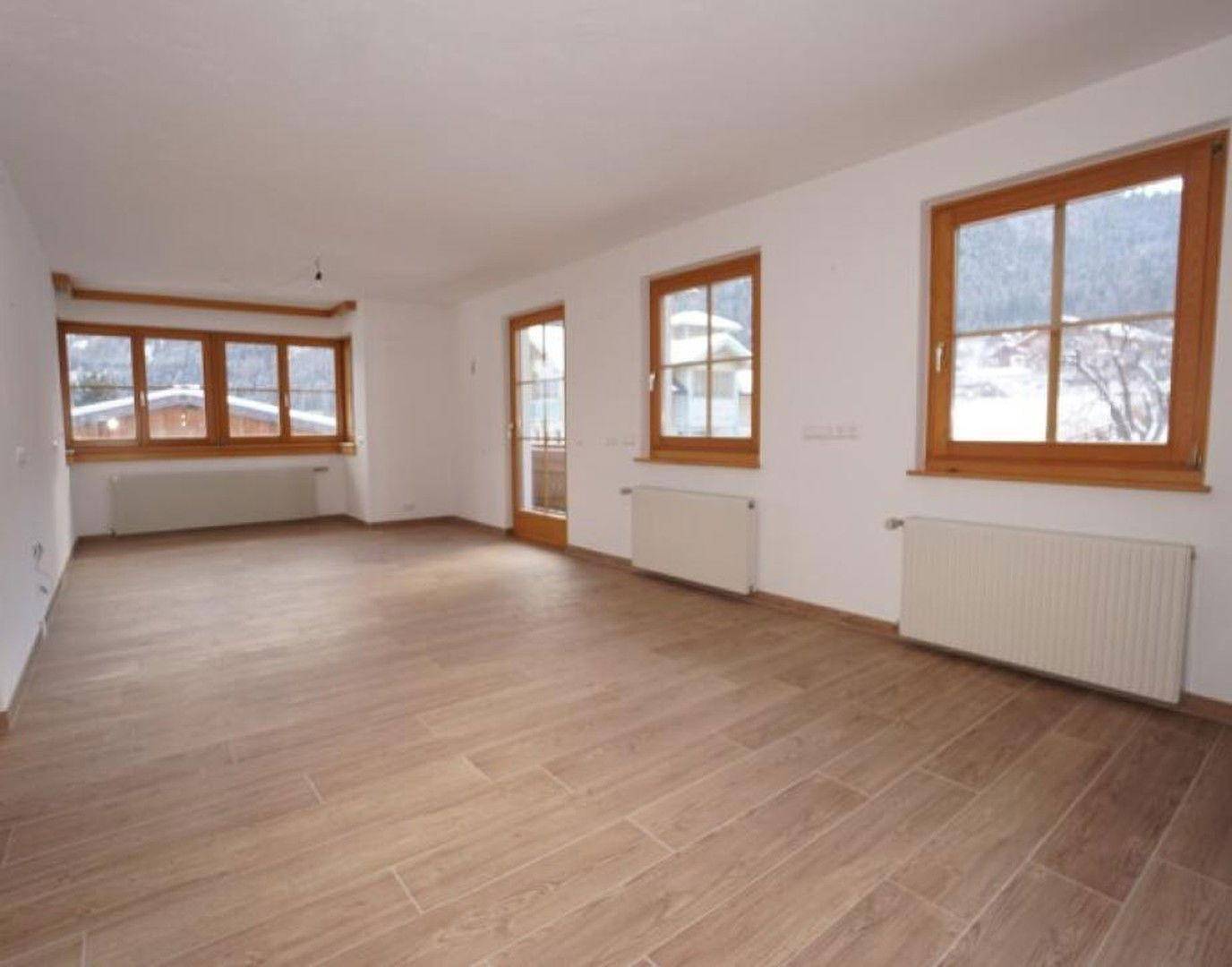 Helles, leeres Wohnzimmer mit großen Fenstern, Fliesenboden und Blick auf die verschneite Landschaft.
