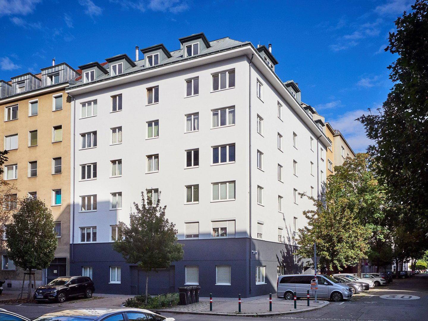 Modernisiertes Mehrfamilienhaus mit weißer Fassade, dunklem Sockel und Mansarddach in städtischer Umgebung.