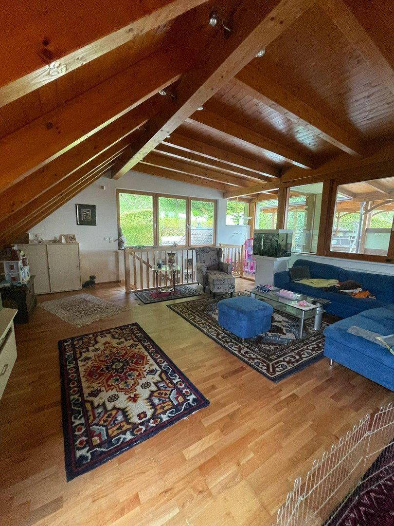 Gemütliches Wohnzimmer im Dachgeschoss mit sichtbaren Holzbalken und großen Fenstern mit Blick ins Grüne.