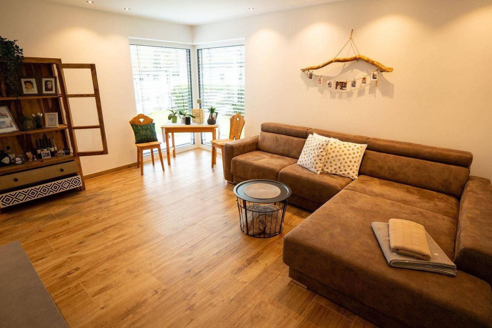 Gemütliches Wohnzimmer mit braunem Ecksofa, Holzboden und großen Fenstern für viel Licht.