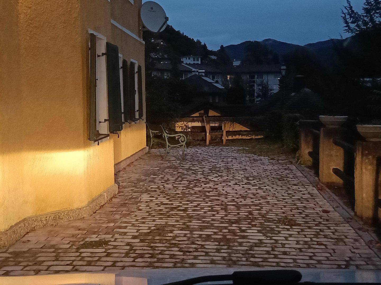 Gepflasterter Außenbereich neben dem Haus in der Abenddämmerung, umgeben von Bäumen.