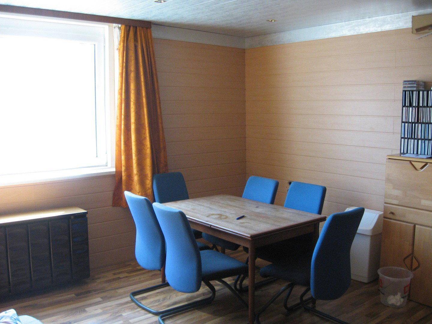 Esszimmer mit Holztisch, blauen Stühlen und holzverkleideten Wänden, gut beleuchtet durch ein Fenster.