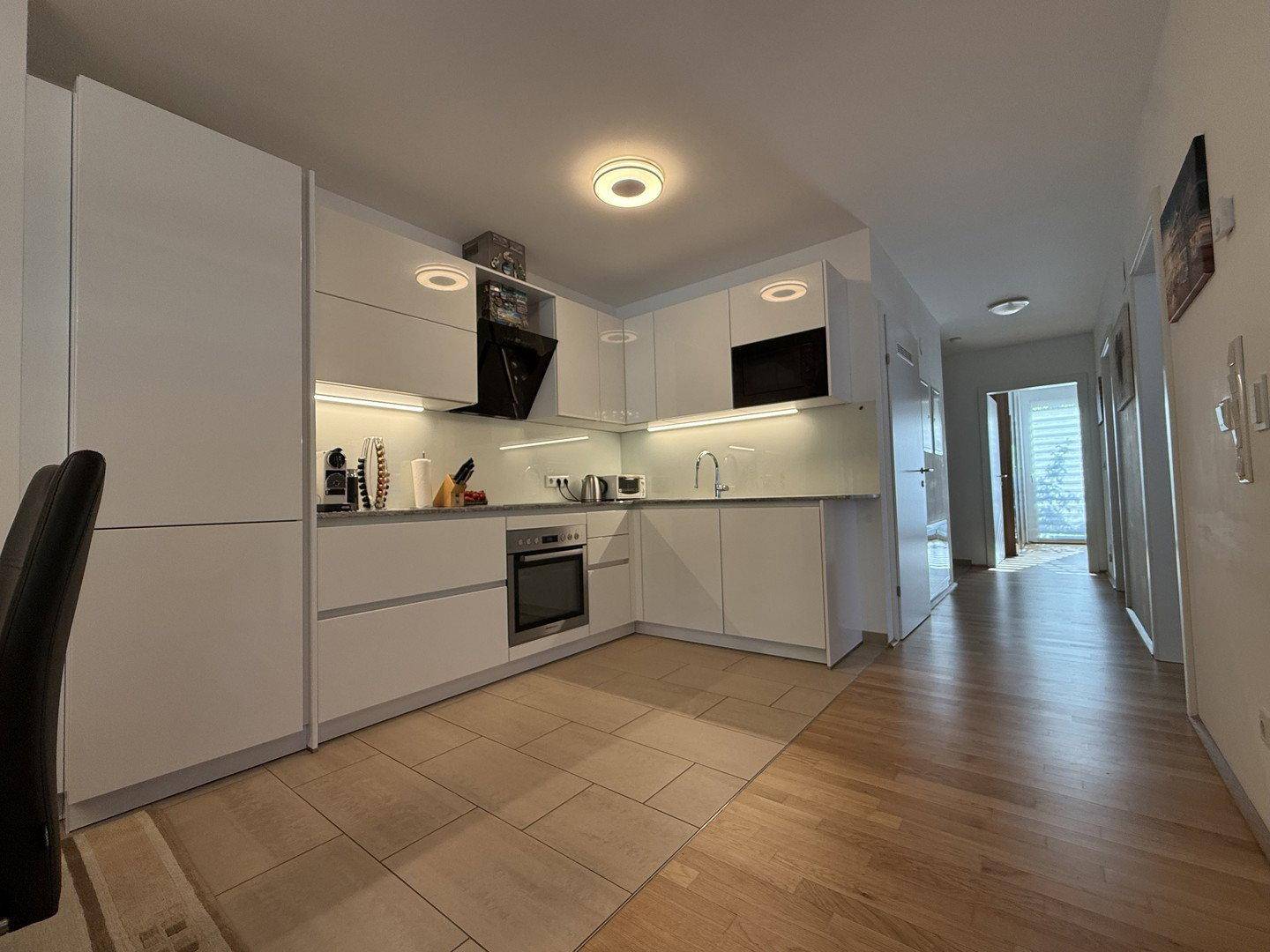 Moderne weiße Küche mit Einbaugeräten, Fliesenboden und Holzboden im angrenzenden Flur.