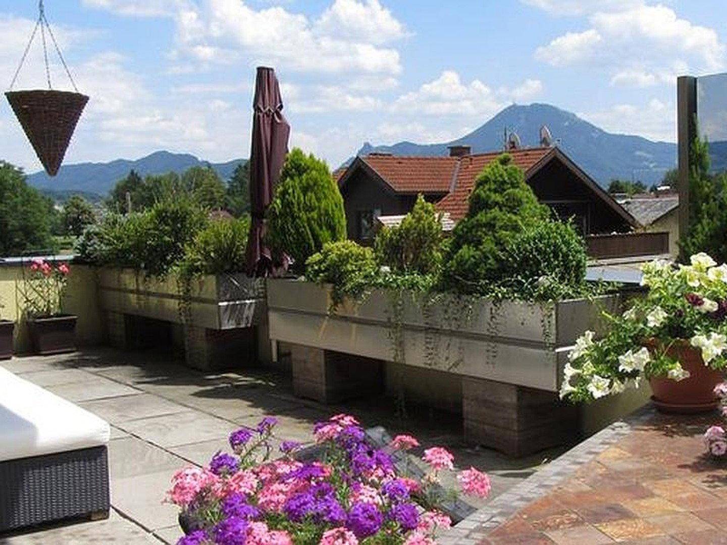 Große Dachterrasse mit Blumenbeeten, Sonnenschirm und Bergblick.