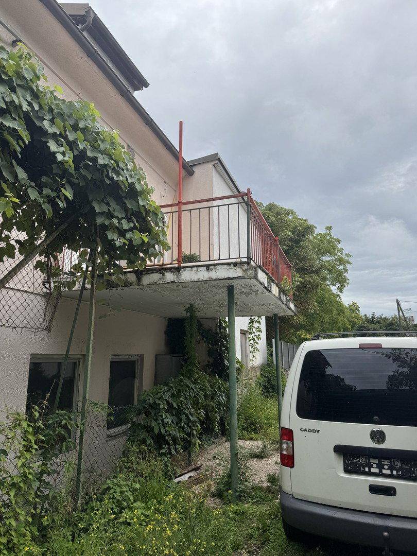 Außenansicht des Gebäudes mit einem älteren Balkon und üppiger Vegetation.