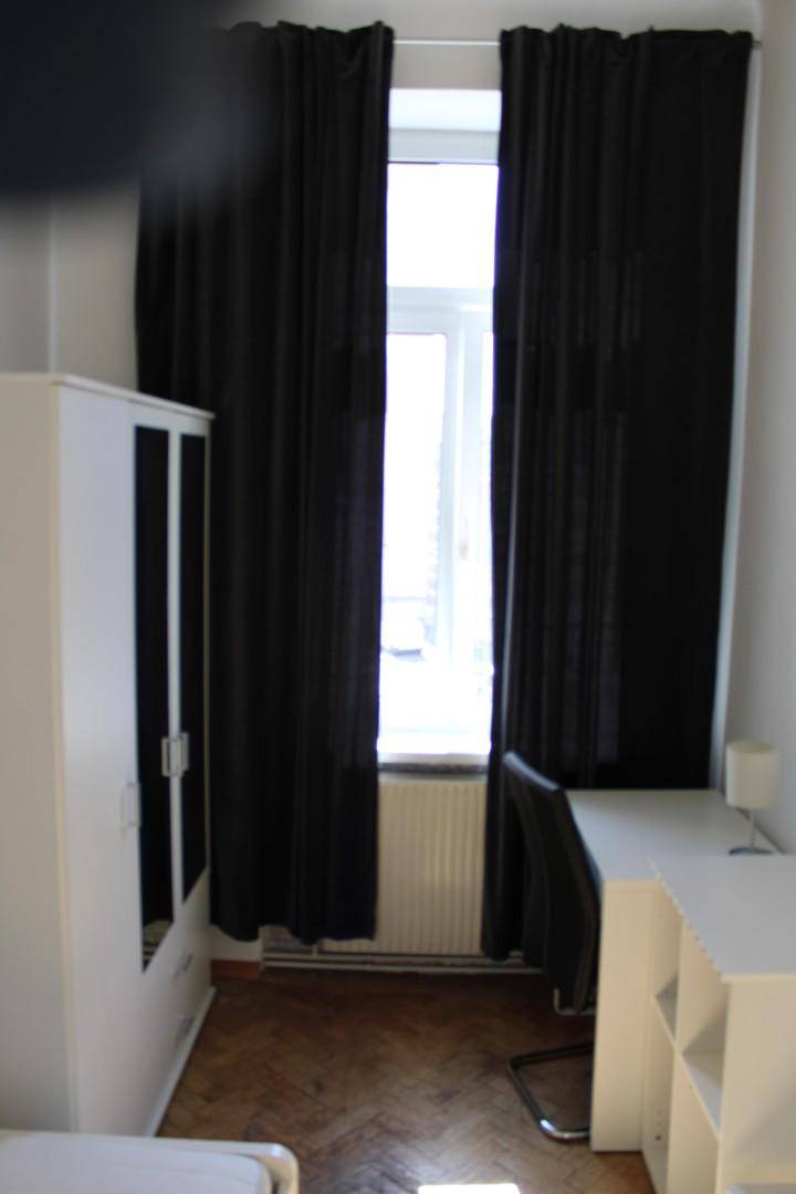 Schlafzimmer mit Schreibtisch, Stuhl und einem Fenster, das von dunklen Vorhängen eingerahmt wird.