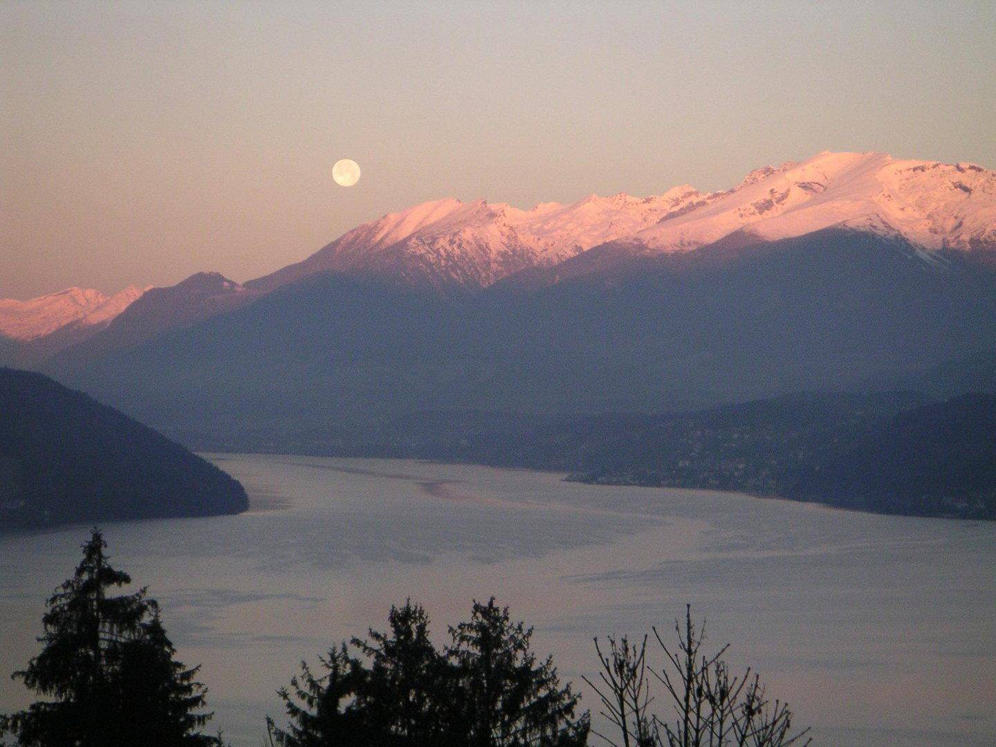 Malerischer Sonnenuntergang über einem See mit schneebedeckten Bergen im Hintergrund und Vollmond.