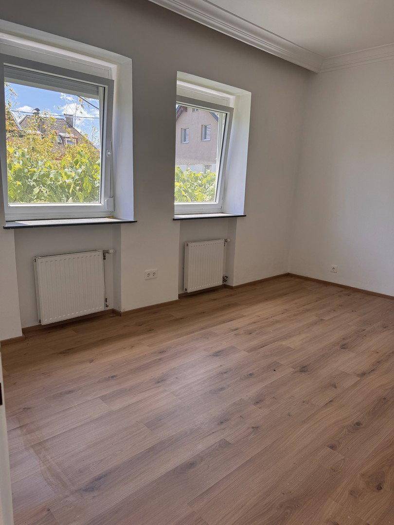 Lichtdurchfluteter Raum mit Holzboden und zwei Fenstern mit Blick ins Grüne.
