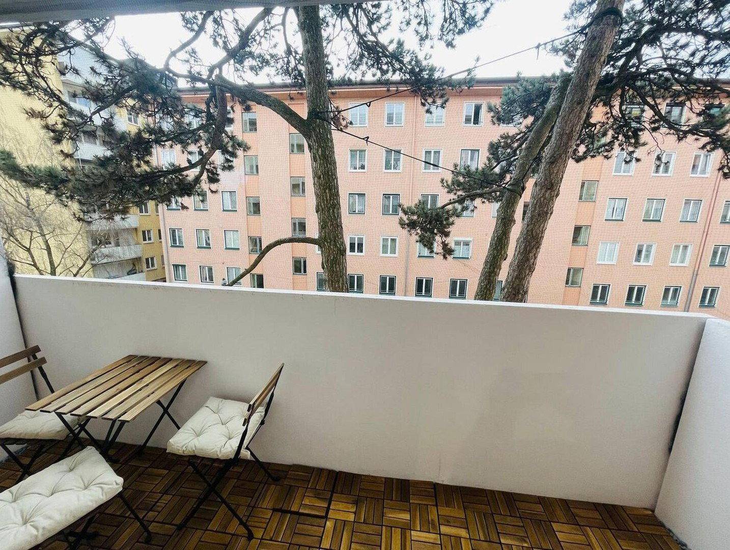 Kleiner Balkon mit Holzboden und Sitzgelegenheiten, umgeben von Bäumen und Gebäuden.