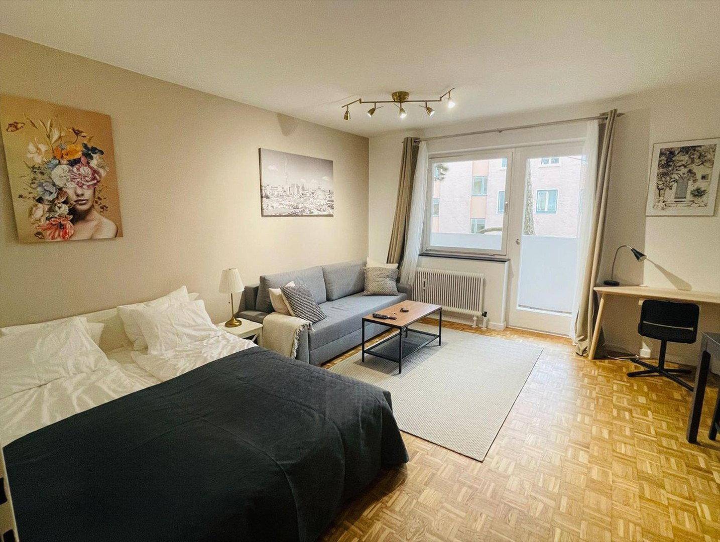 Wohnzimmer mit Schlafbereich, Sofa, Schreibtisch und Balkonzugang.