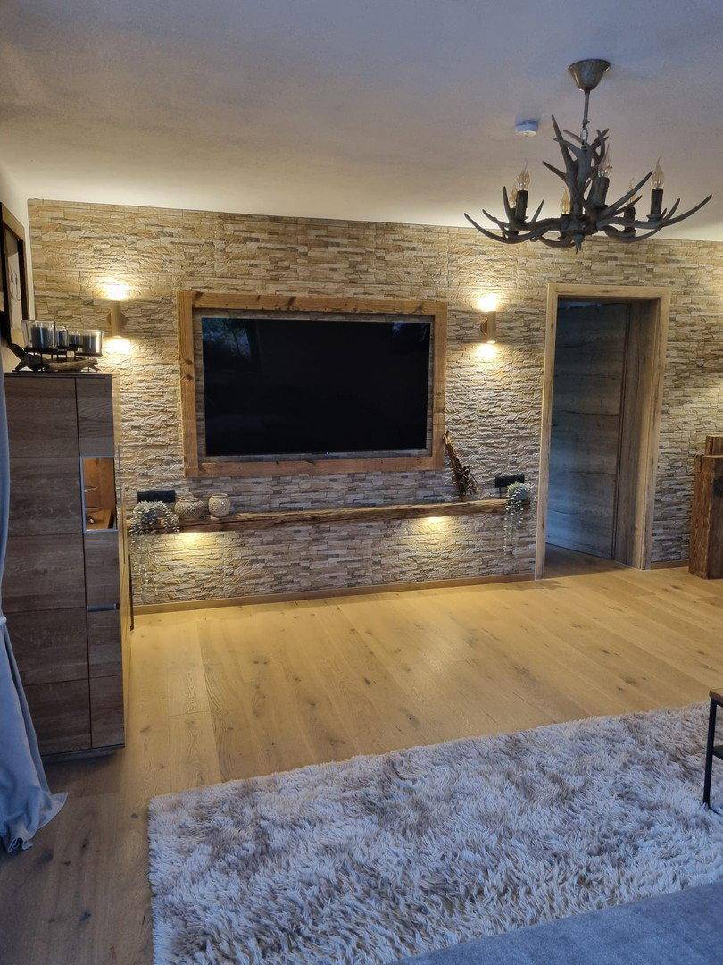Wohnzimmer mit Steinwand, großem Fernseher und Geweihleuchter, Holzboden.