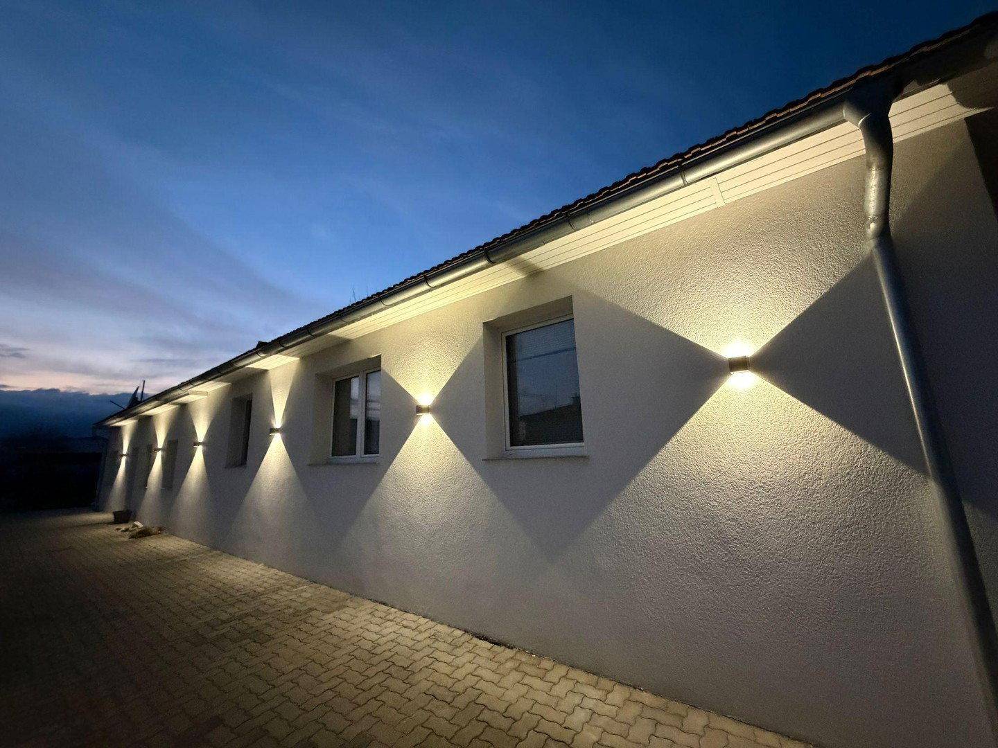 Fassade des Gebäudes bei Dämmerung mit stilvoller Wandbeleuchtung und gepflastertem Weg.