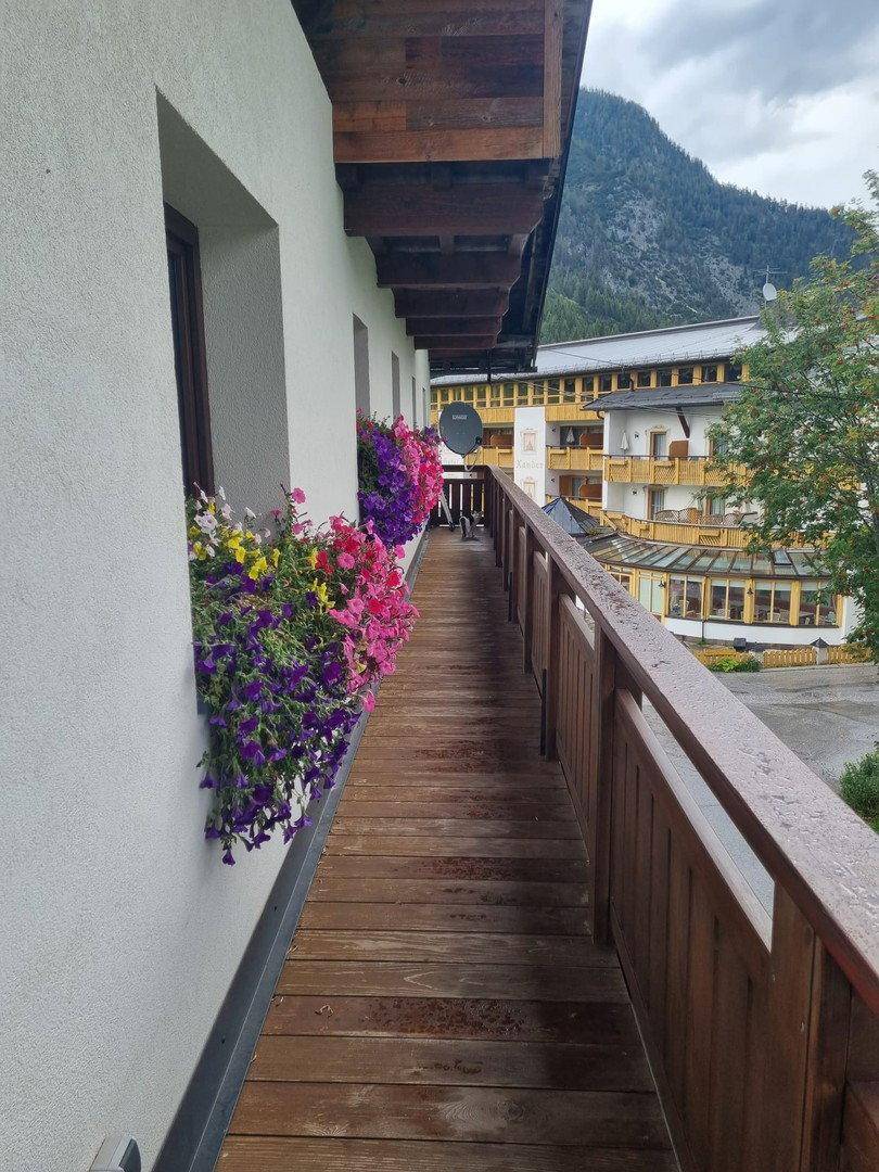 Langer Holzbalkon mit bunten Blumenkästen und Blick auf die umliegenden Gebäude und Berge.