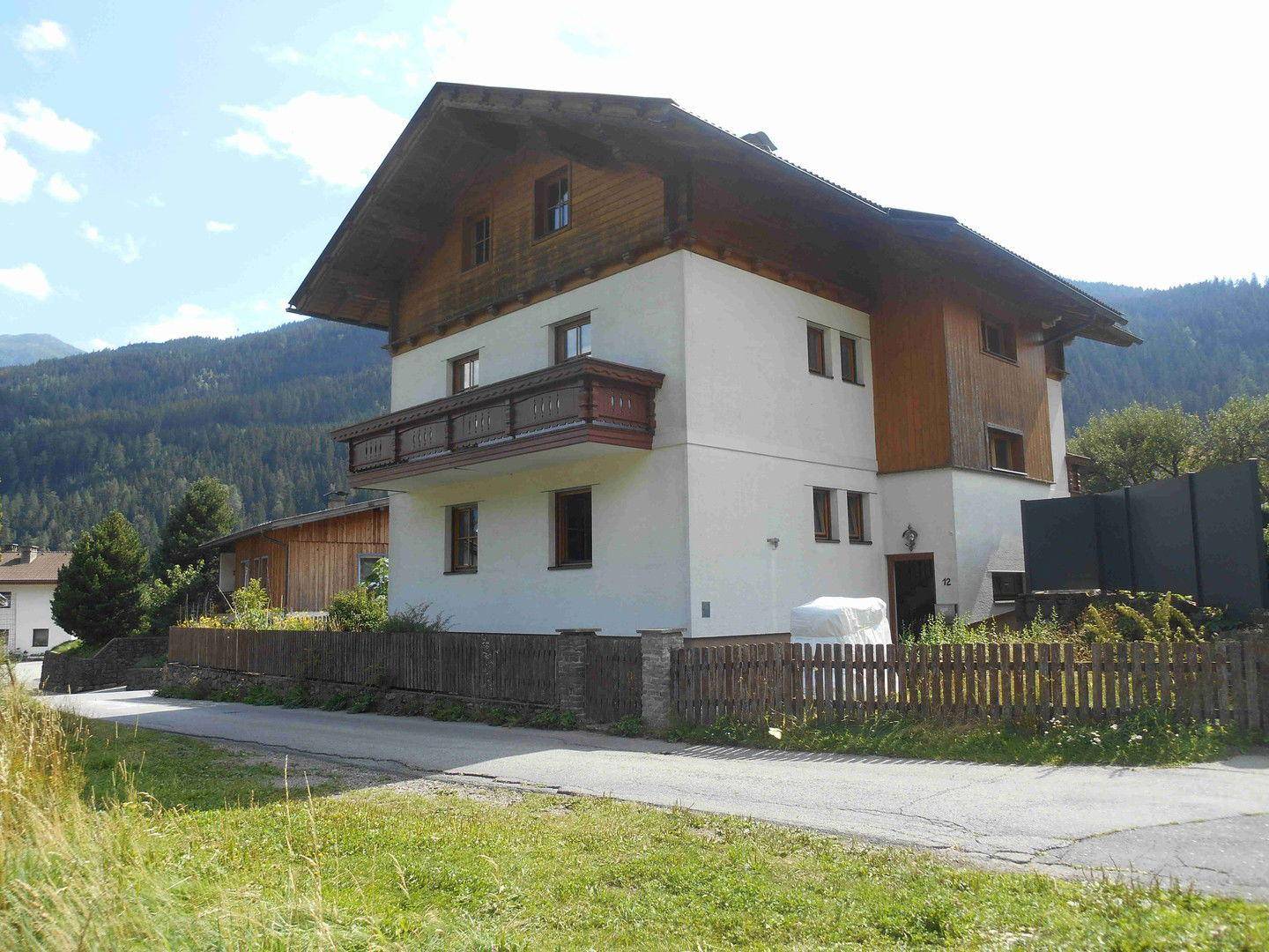 Ansicht des traditionellen Hauses mit Holzbalkon und weißer Fassade, umgeben von grüner Landschaft und Bergen.