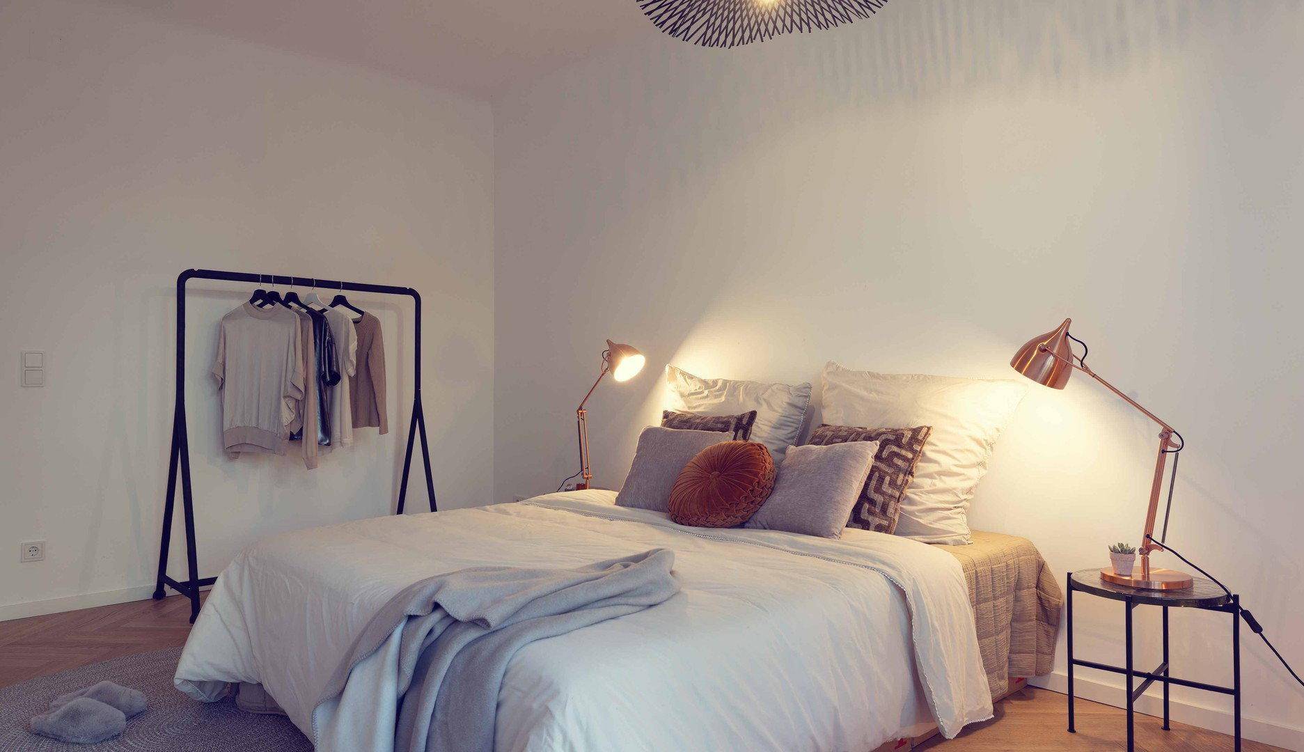 Schlafzimmer mit Doppelbett, Kleiderstange und warmem Licht der Nachttischlampen.