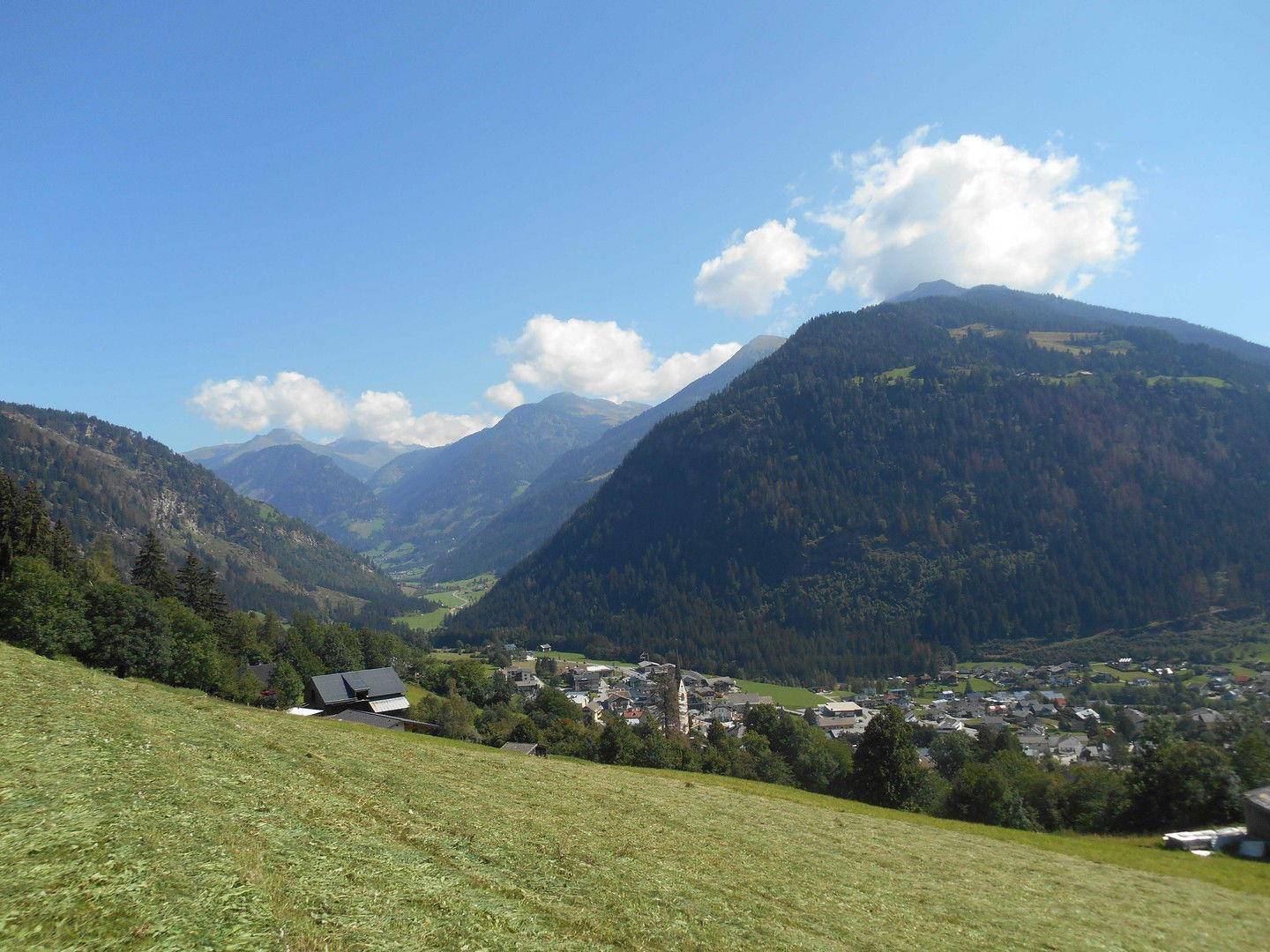 Malerischer Panoramablick auf ein alpines Tal mit Dorf, grünen Wiesen und hohen Bergen.