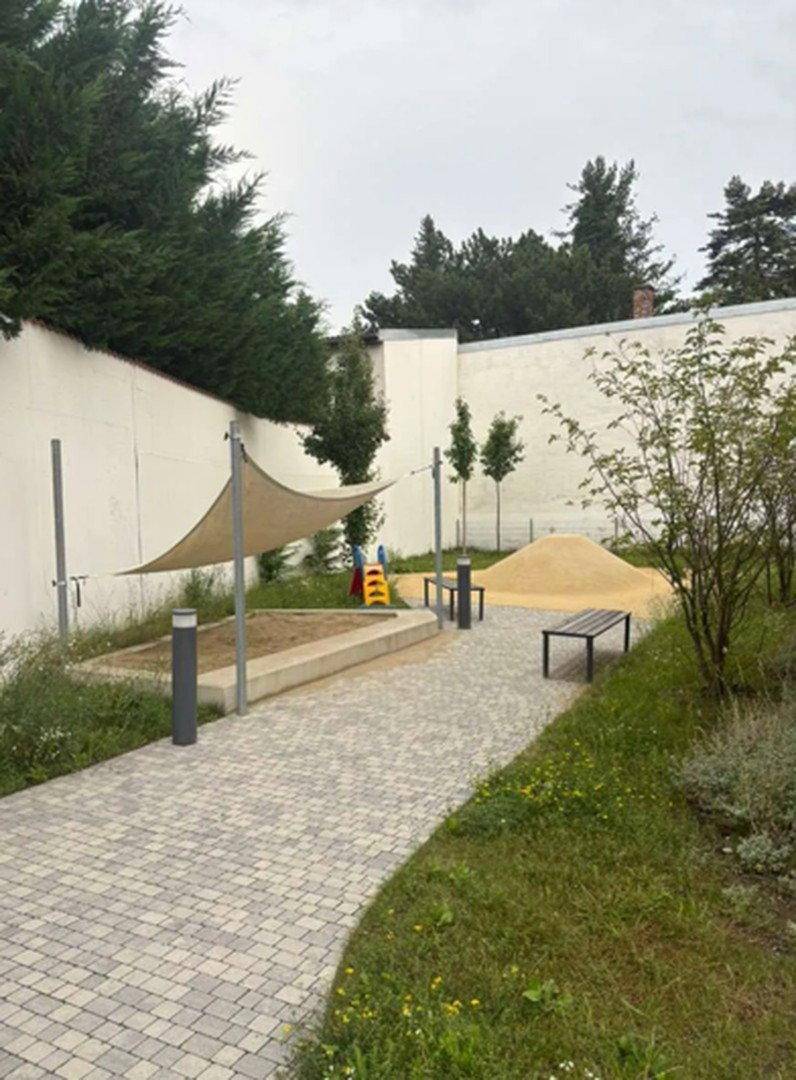 Kinderspielplatz im Freien mit Sandkasten, Rutsche und Sitzbänken unter einem Sonnensegel.