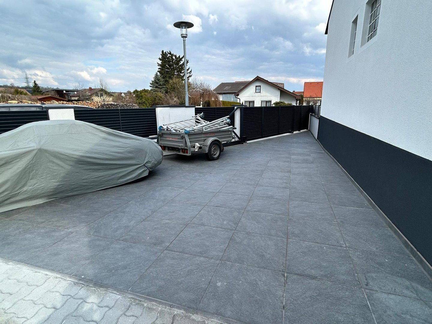 Gepflasterter Parkplatz mit abgedecktem Auto und Anhänger neben einem schwarzen Zaun.