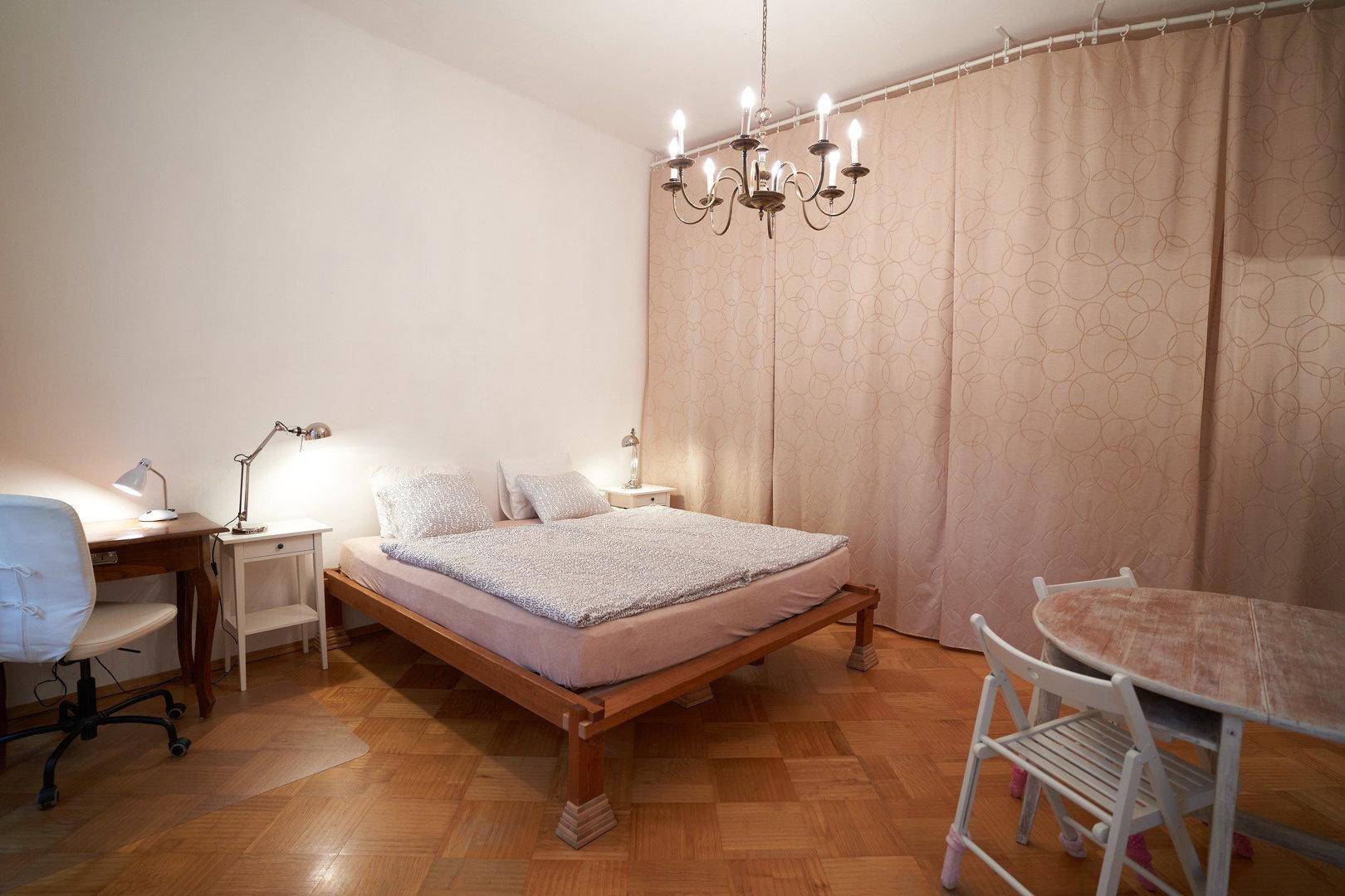 Gemütliches Schlafzimmer mit Holzbett, Schreibtisch und einem kleinen Tischbereich, beleuchtet von einem Kronleuchter.