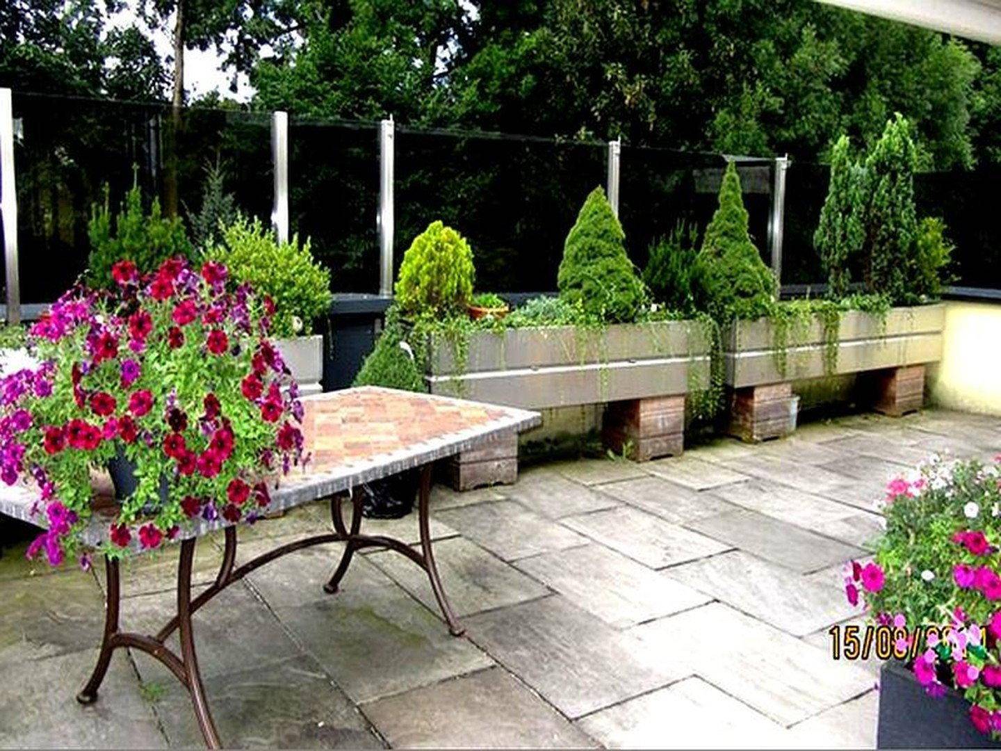 Große gepflasterte Terrasse mit üppigen Blumenbeeten und einem Tisch, umgeben von viel Grün.