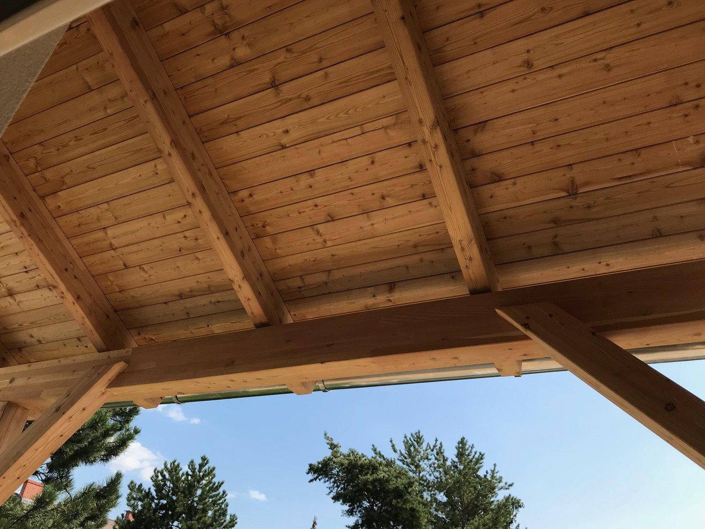 Blick auf die robuste Holzdeckenkonstruktion eines überdachten Balkons unter blauem Himmel.