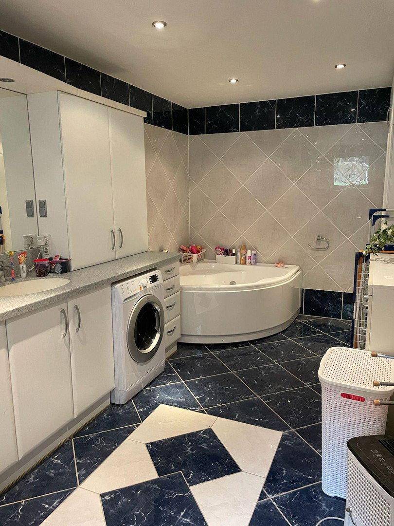 Geräumiges Badezimmer mit Eckbadewanne, Waschmaschine und blauen Fliesen im 80er-Jahre-Stil.