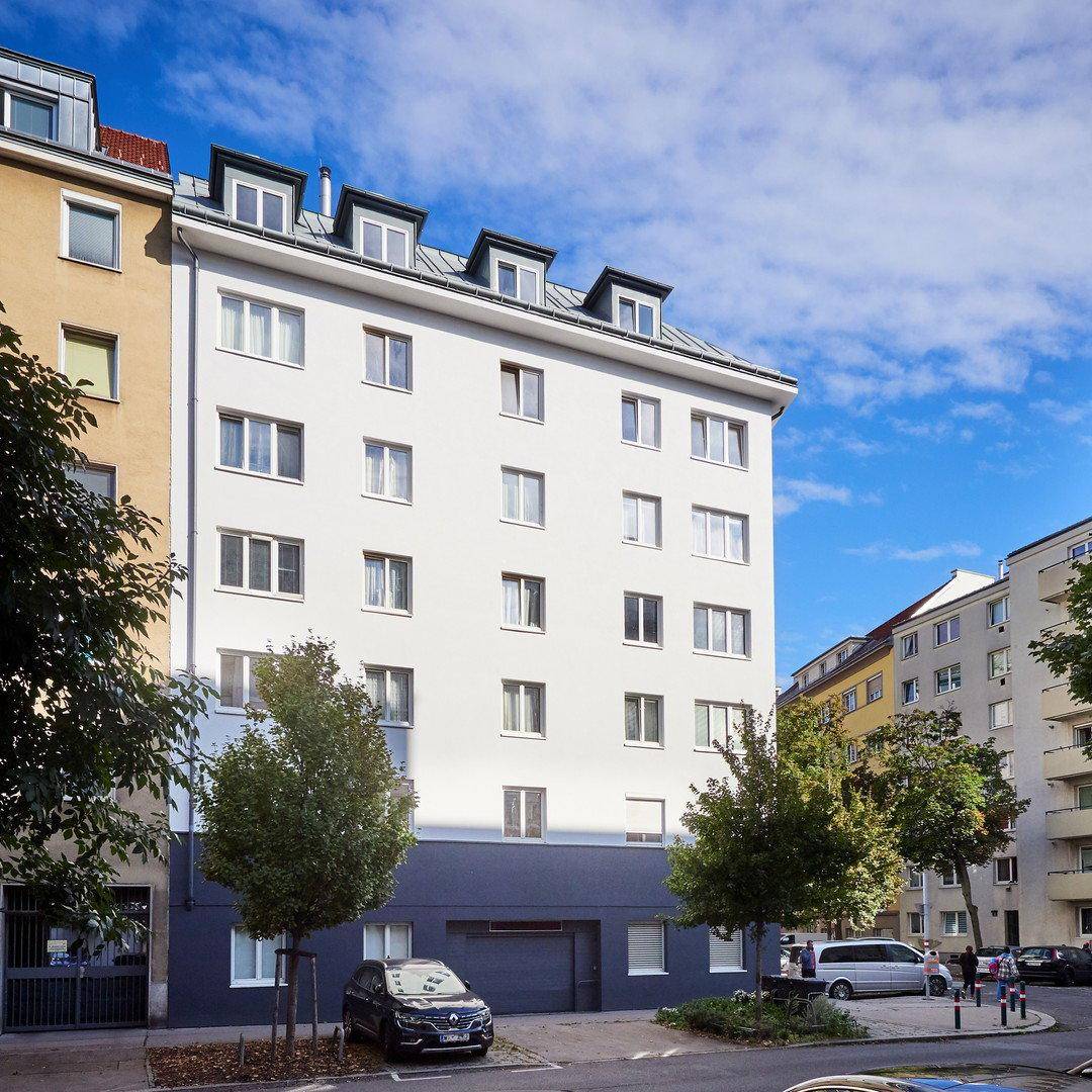 Ansicht eines sanierten Wohngebäudes mit heller Fassade und vielen Fenstern, umgeben von Bäumen und parkenden Autos.