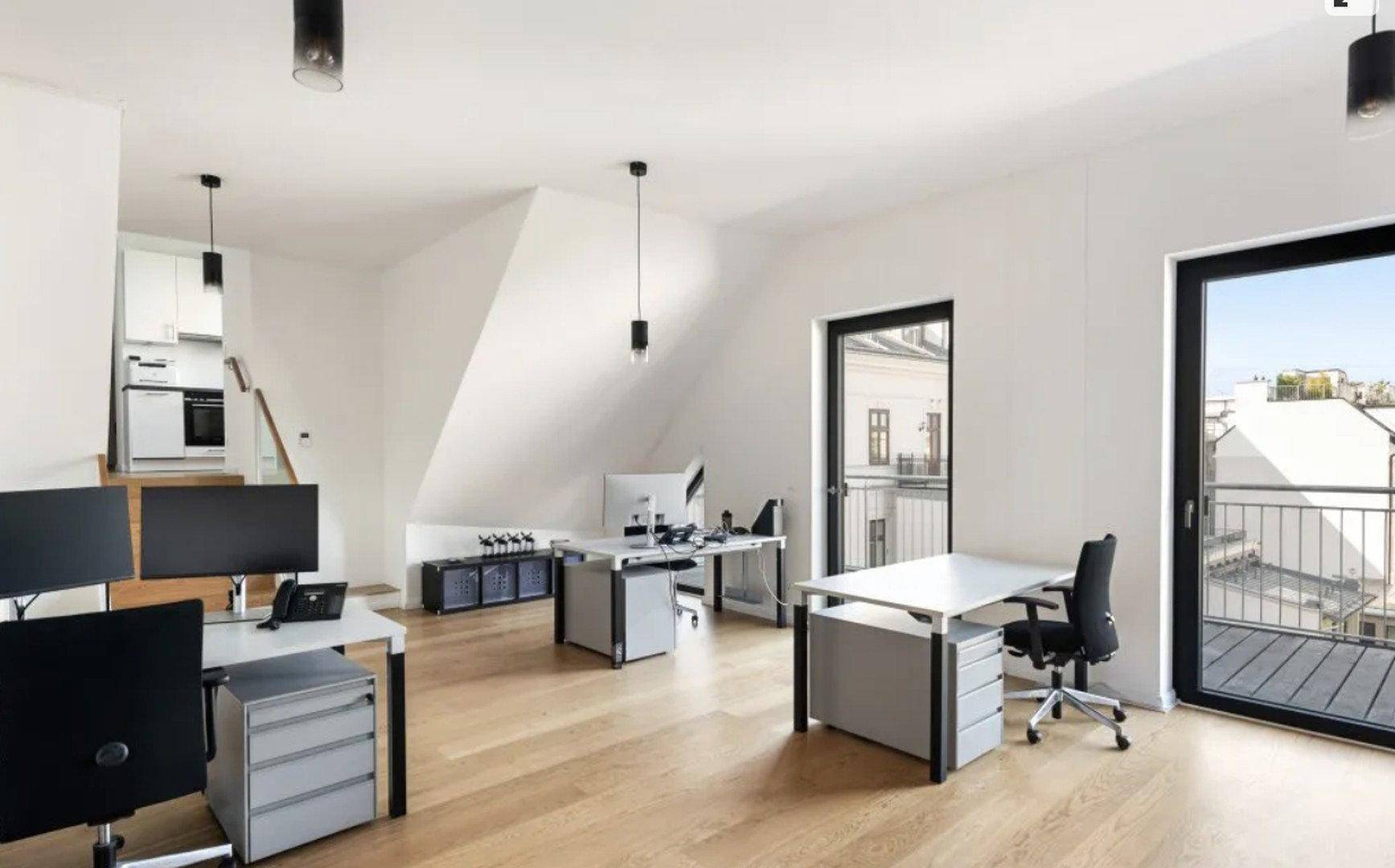 Helles, offenes Büro im Dachgeschoss mit modernen Arbeitsplätzen, Holzboden und Zugang zu einem Balkon.