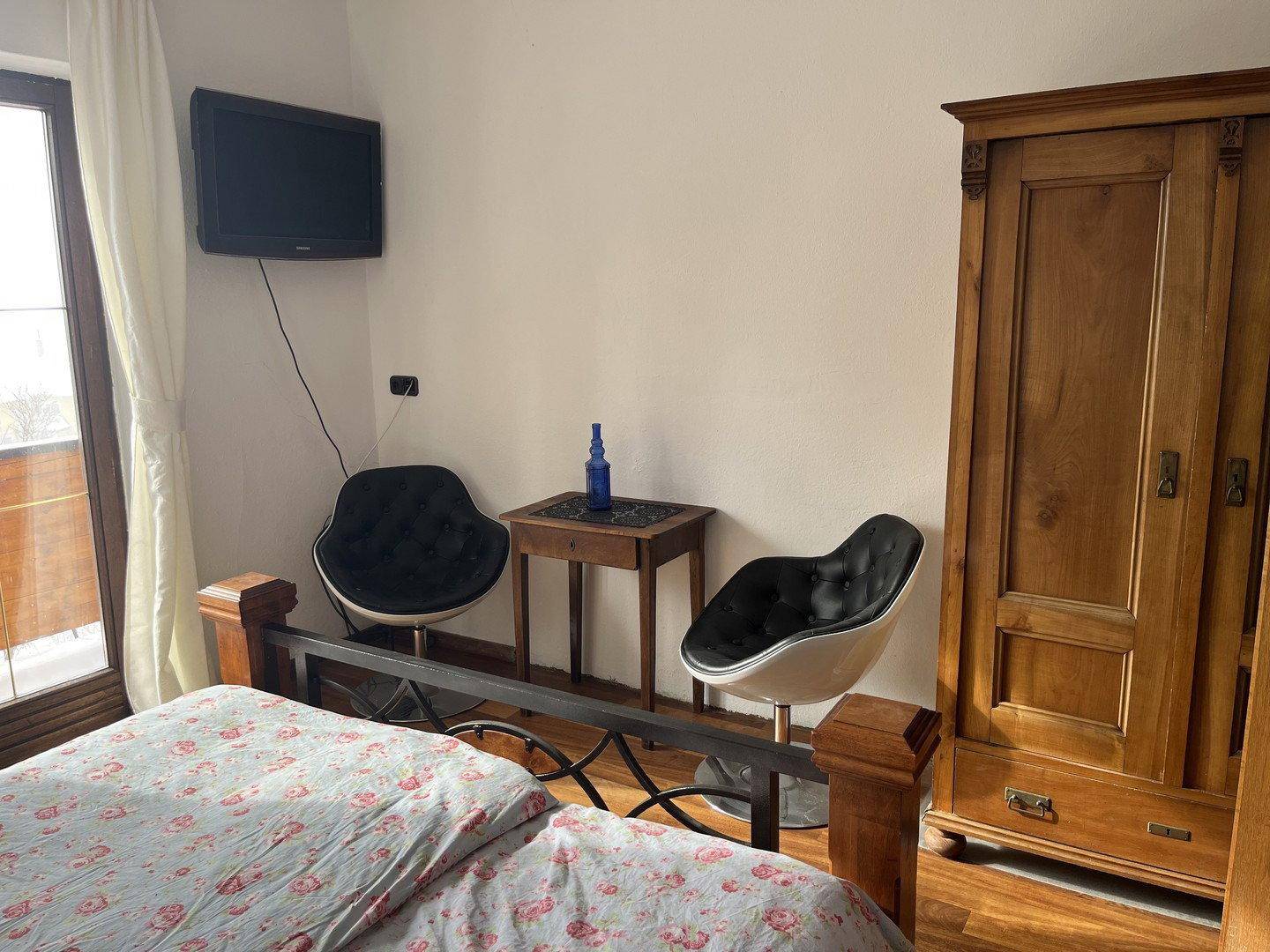 Schlafzimmer mit Holzbett, Kleiderschrank, Fernseher und zwei modernen Stühlen, mit Balkonzugang.
