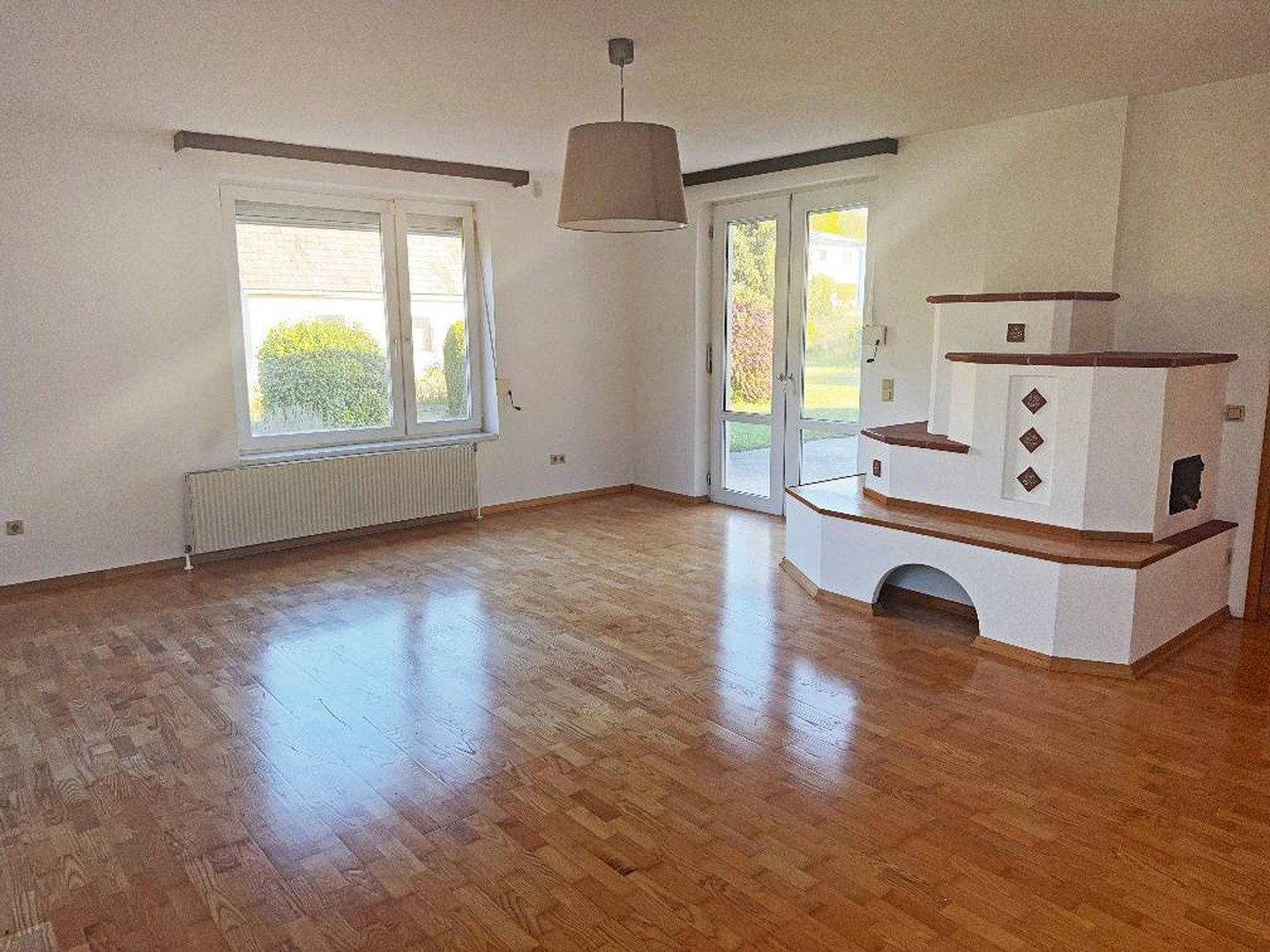 Geräumiges Wohnzimmer mit Kachelofen, Holzfußboden und Zugang zum Garten.